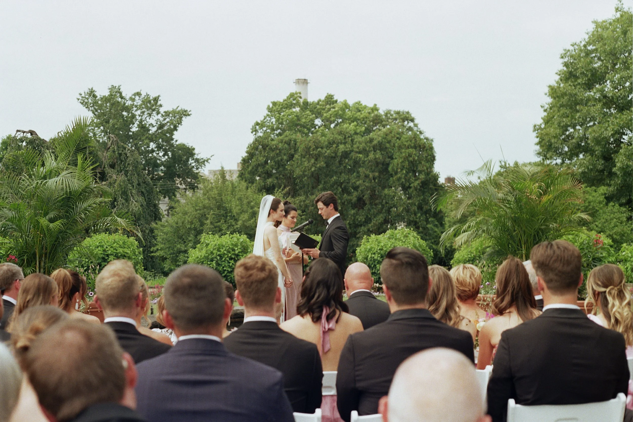 A wedding ceremony with a bride and groom exchanging vows outdoors among guests with lush green trees in the background.
