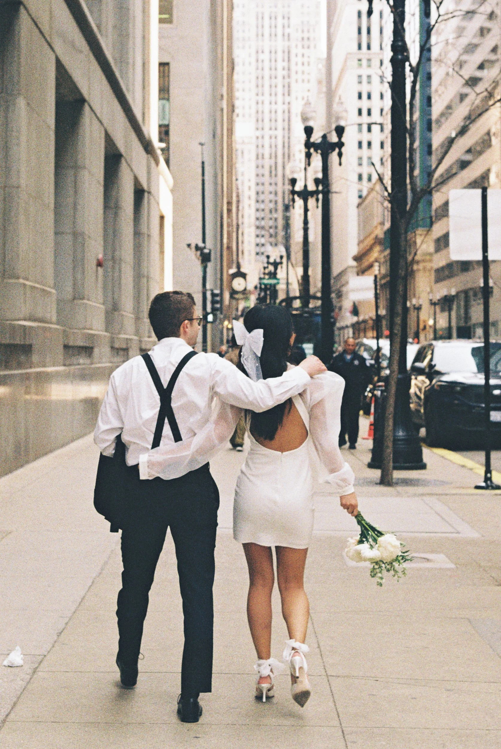 A couple walking down a city street, with the man placing his hand on the woman's shoulder. The woman is dressed in a white dress with an open back, holding a bouquet of white flowers, and wearing high heels with ribbon accents. The scene is set in a