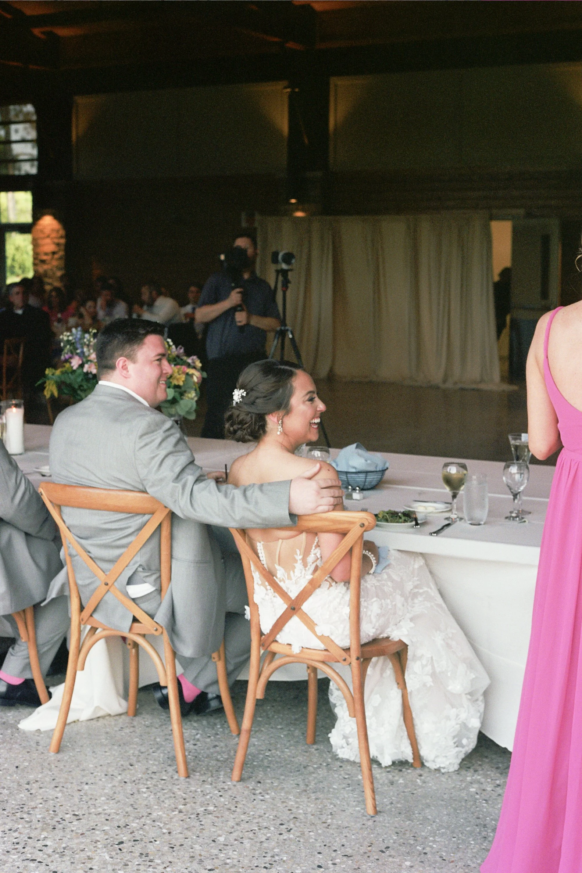 Bride and groom sitting at a wedding reception table, smiling and laughing, with guests in the background and a photographer capturing moments.