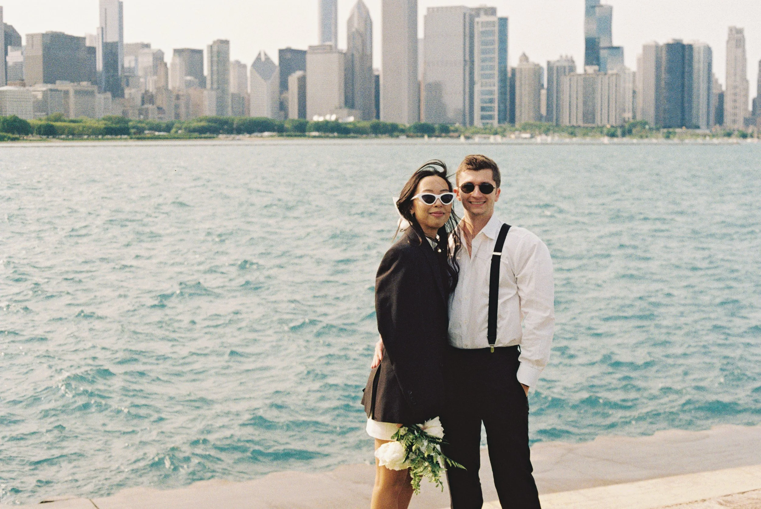 A couple standing by the water with a city skyline in the background, the woman holding a bouquet of flowers.