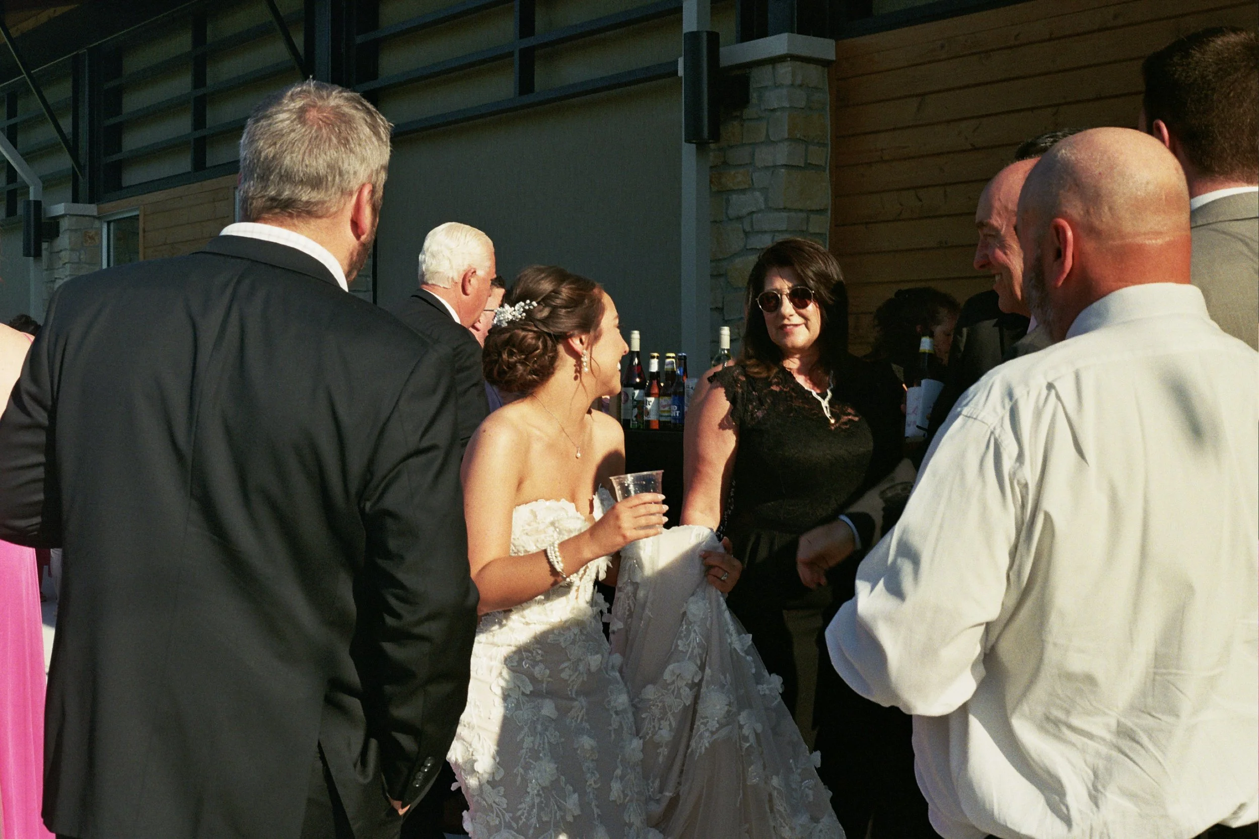 Group of people at a wedding reception, including a bride in a strapless wedding dress holding a drink, talking with guests outdoors in sunny weather.