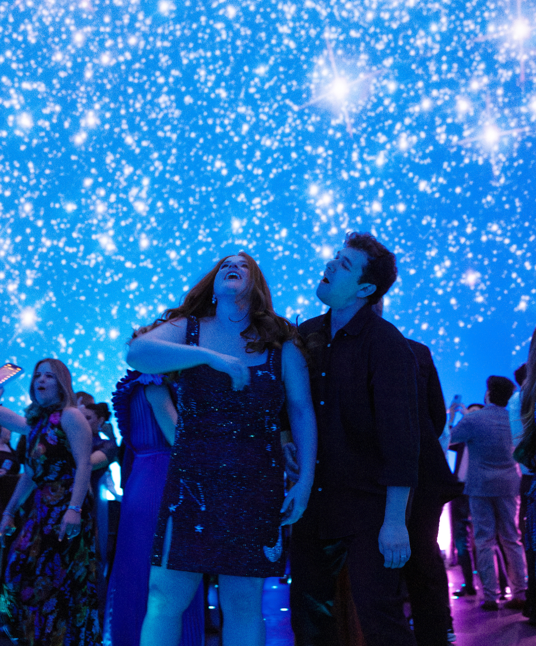 Two people at a party with a starry sky projection on the ceiling, smiling and singing, with other guests in the background.