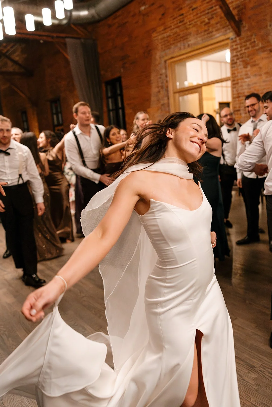 A woman in a white wedding dress dancing joyfully at a wedding reception with guests in the background.
