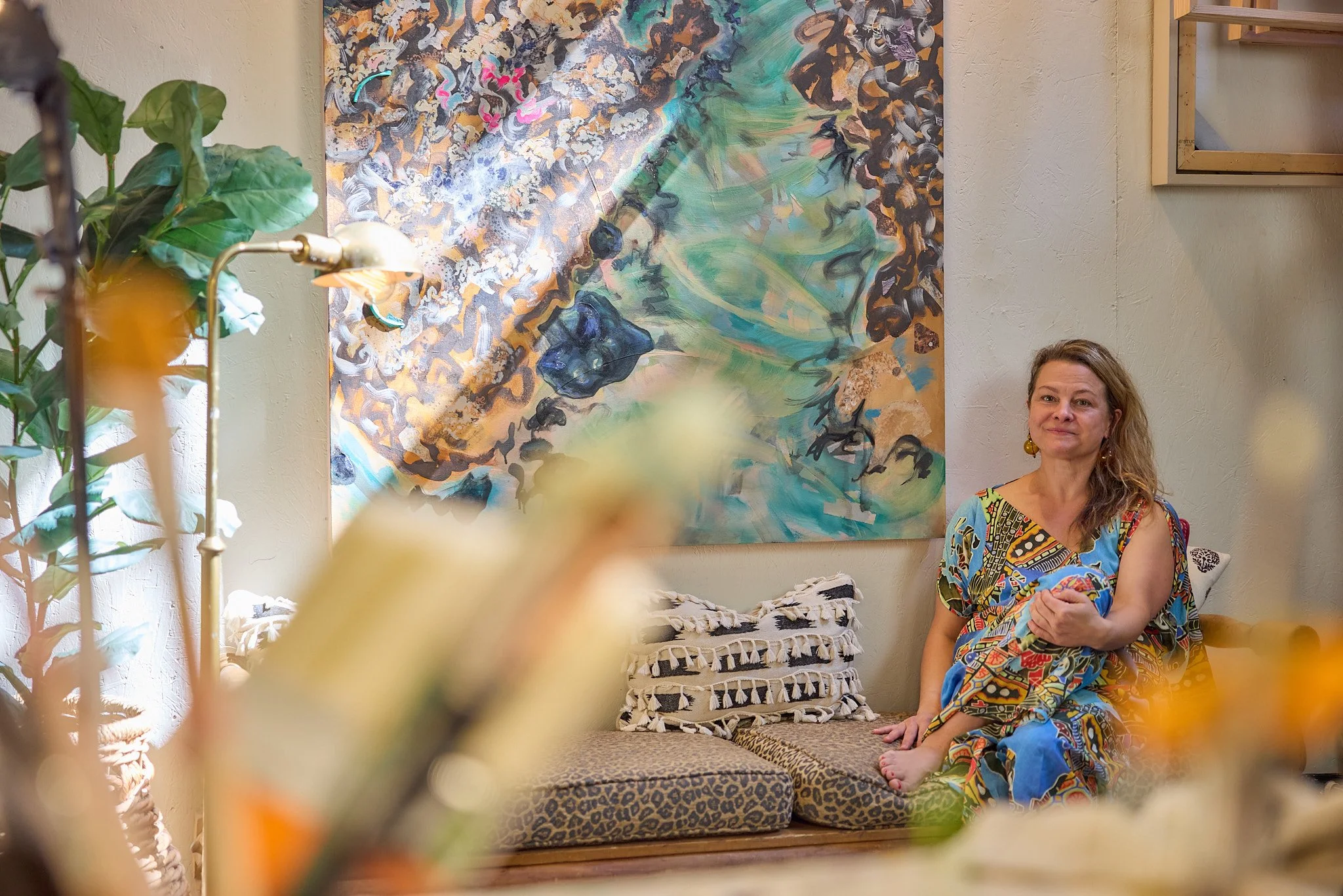 Portrait of Rossi seated on a bench in her studio next to "Coral," 60 x 46." It is a colorful painting. She is wearing a colorful dress of similar colors - blue, green, brown, yellow - and smiling. Sunlight highlights diagonal bands.
