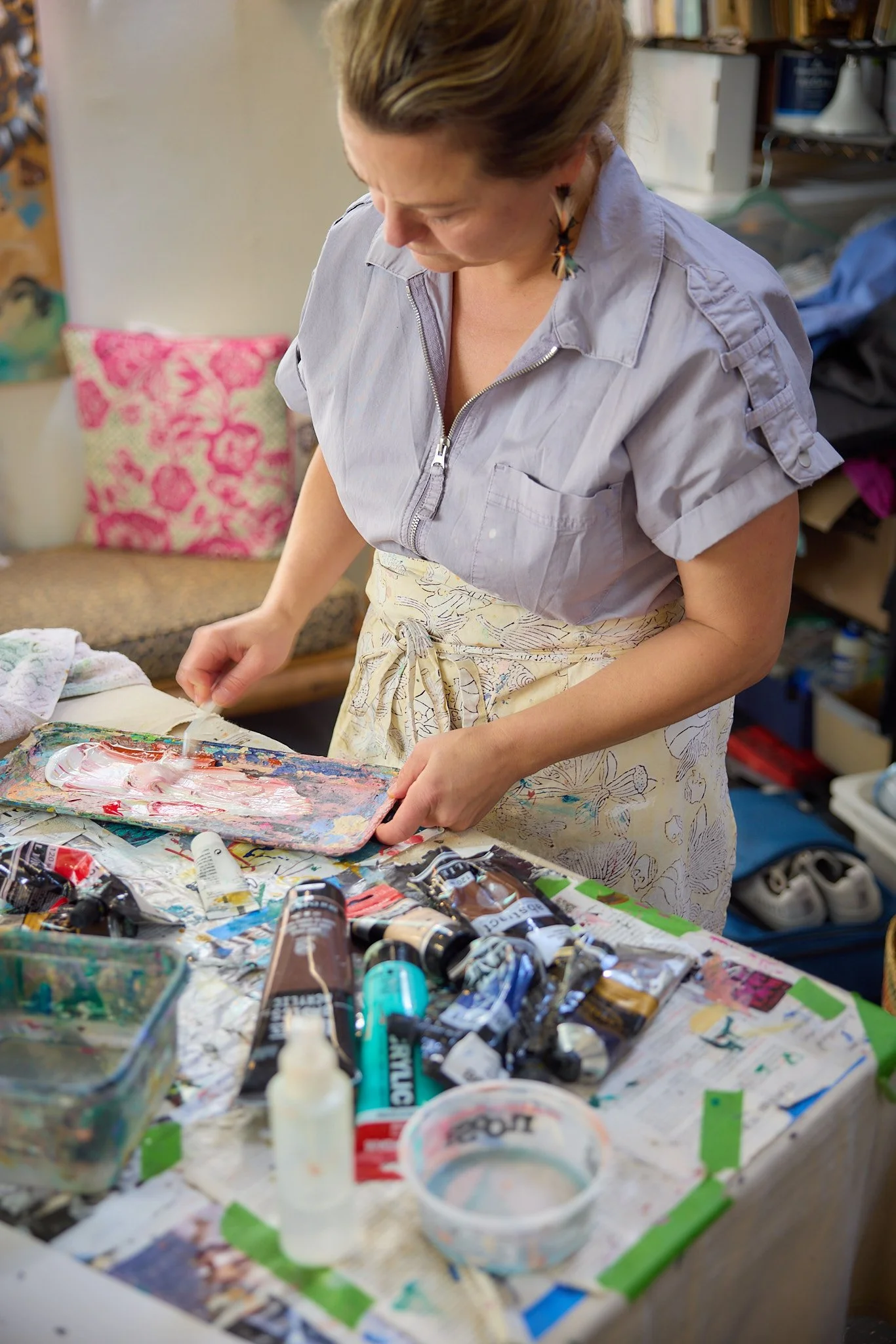The artist Rossi in an apron mixing paint on a palette in her art studio, surrounded by tubes of paint, brushes and art supplies on a table.