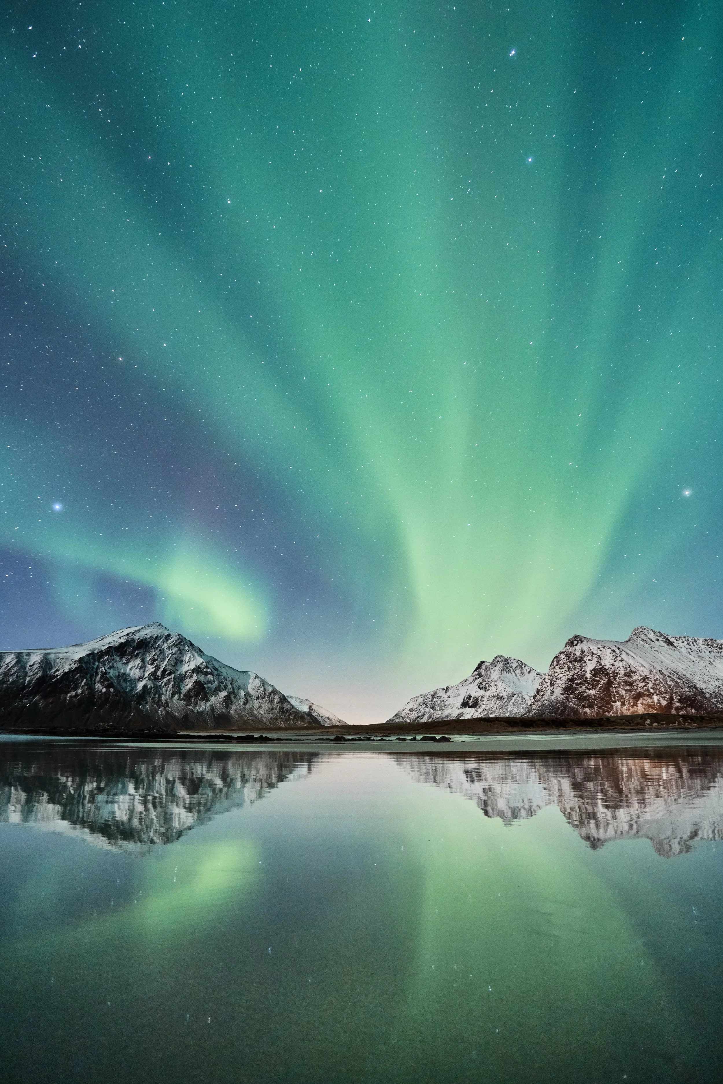 Northern lights over snow-capped mountains reflected in a calm body of water at night.