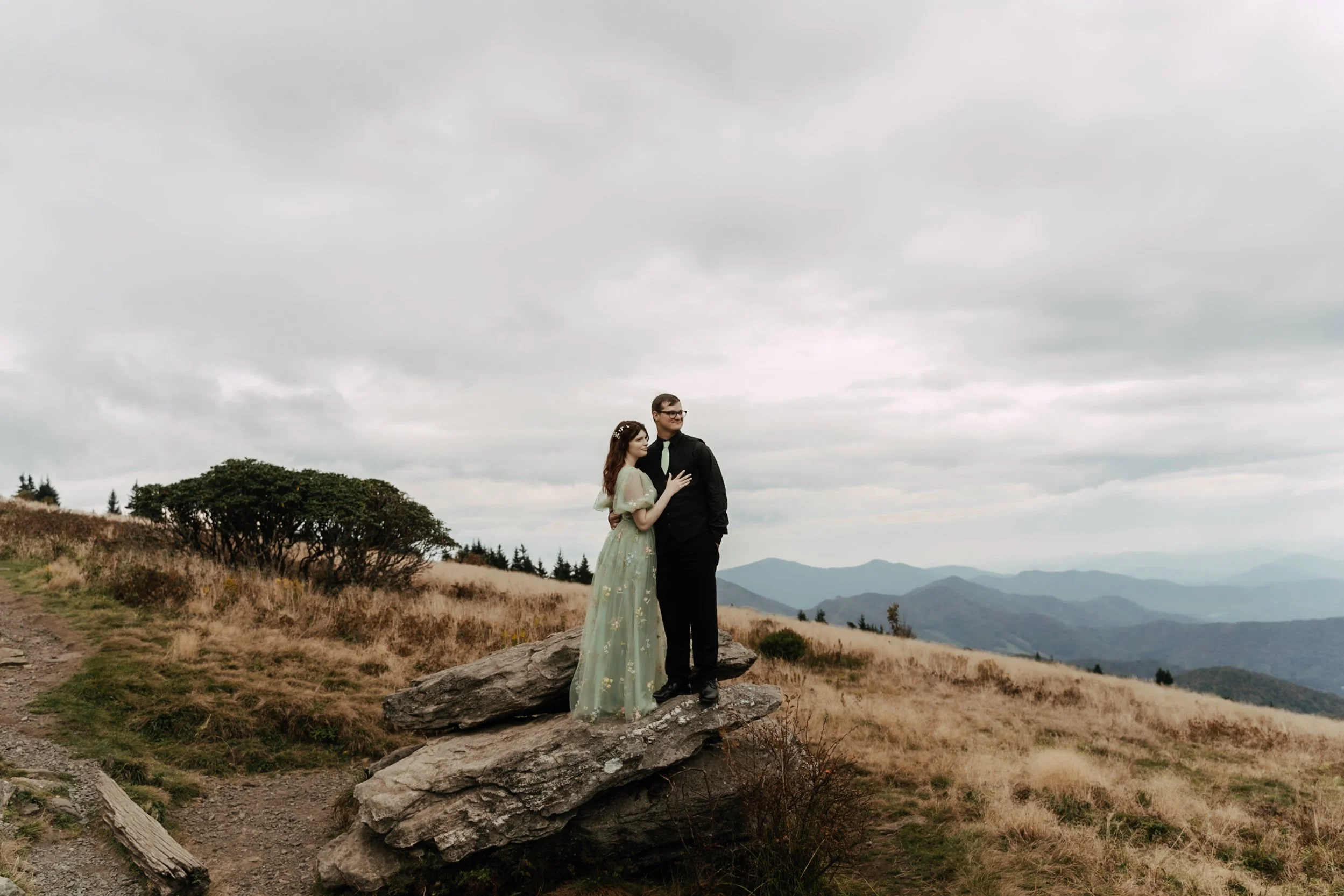 A couple dressed in formal attire standing on a large rock in a mountainous landscape, with rolling mountains and cloudy sky in the background.