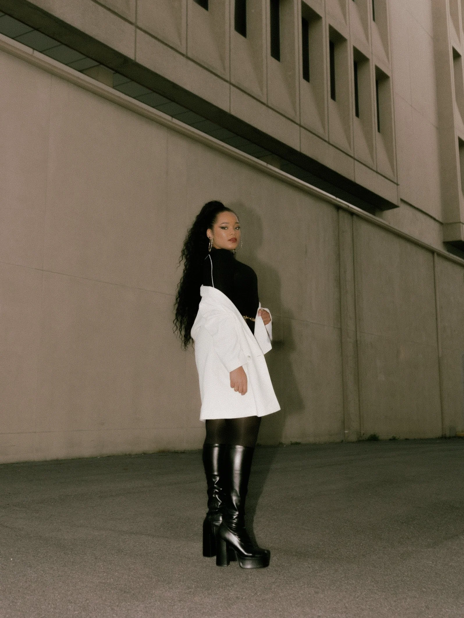 A woman standing on the street at night, wearing a black top, white blazer, and black thigh-high platform boots, with large earrings and long curly hair.