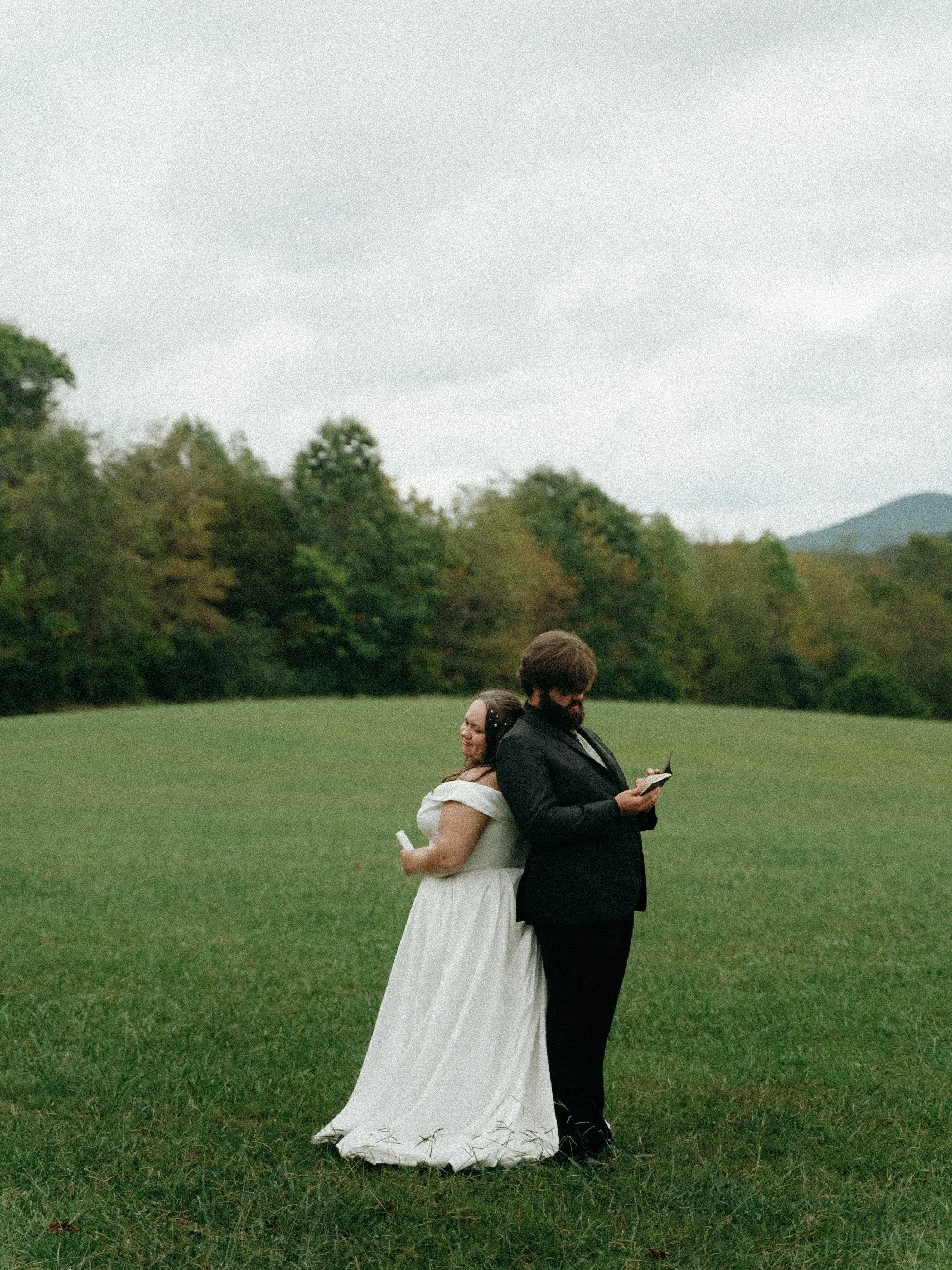 A bride and groom stand back-to-back on a grassy field, holding scrolls, with the bride smiling and the groom reading a book. The background features trees and a cloudy sky.