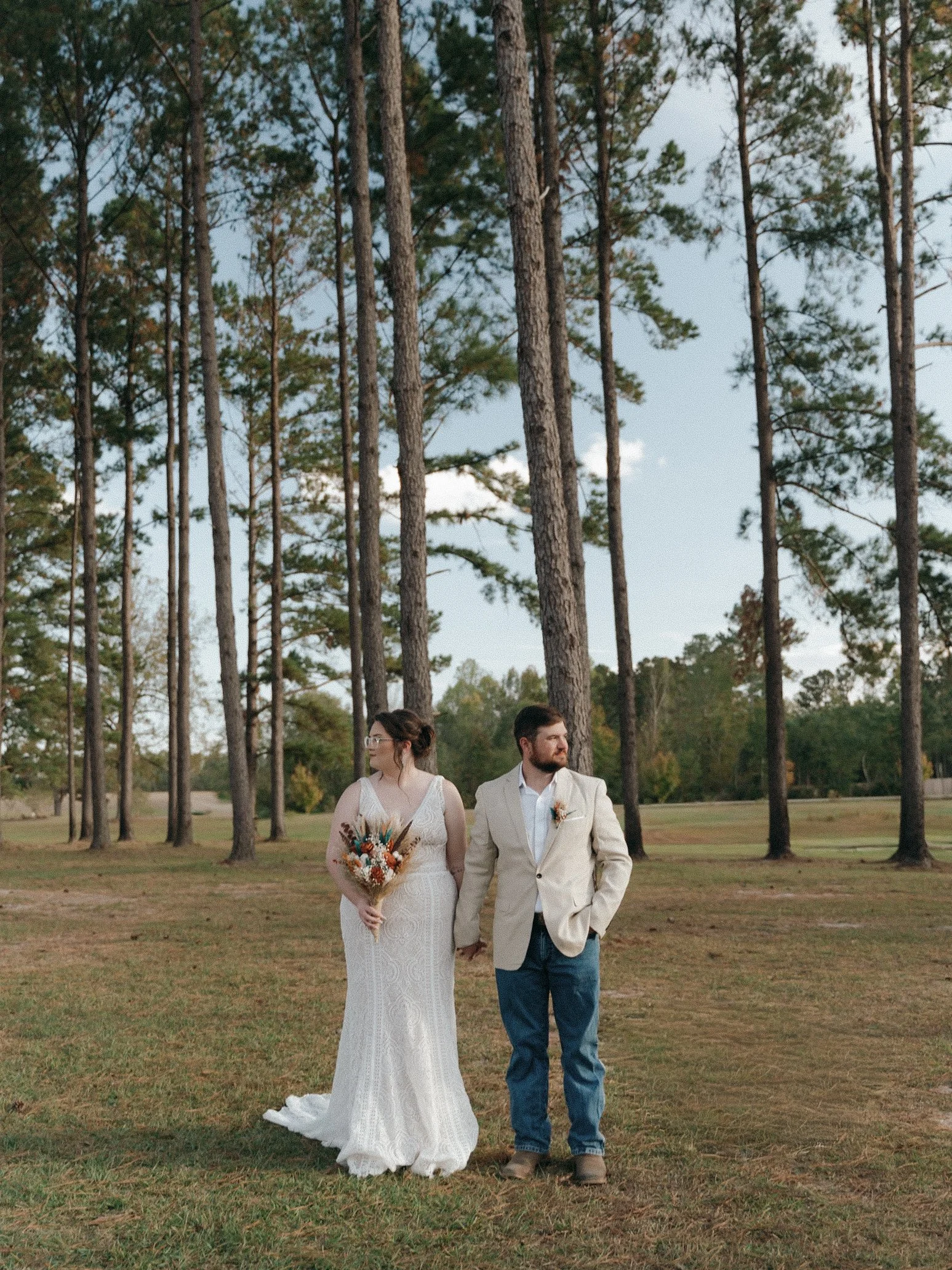 A couple holding hands in a wooded outdoor setting during daylight. The woman is wearing a white lace wedding dress and glasses, holding a bouquet. The man is dressed in a beige blazer, white shirt, and blue jeans.