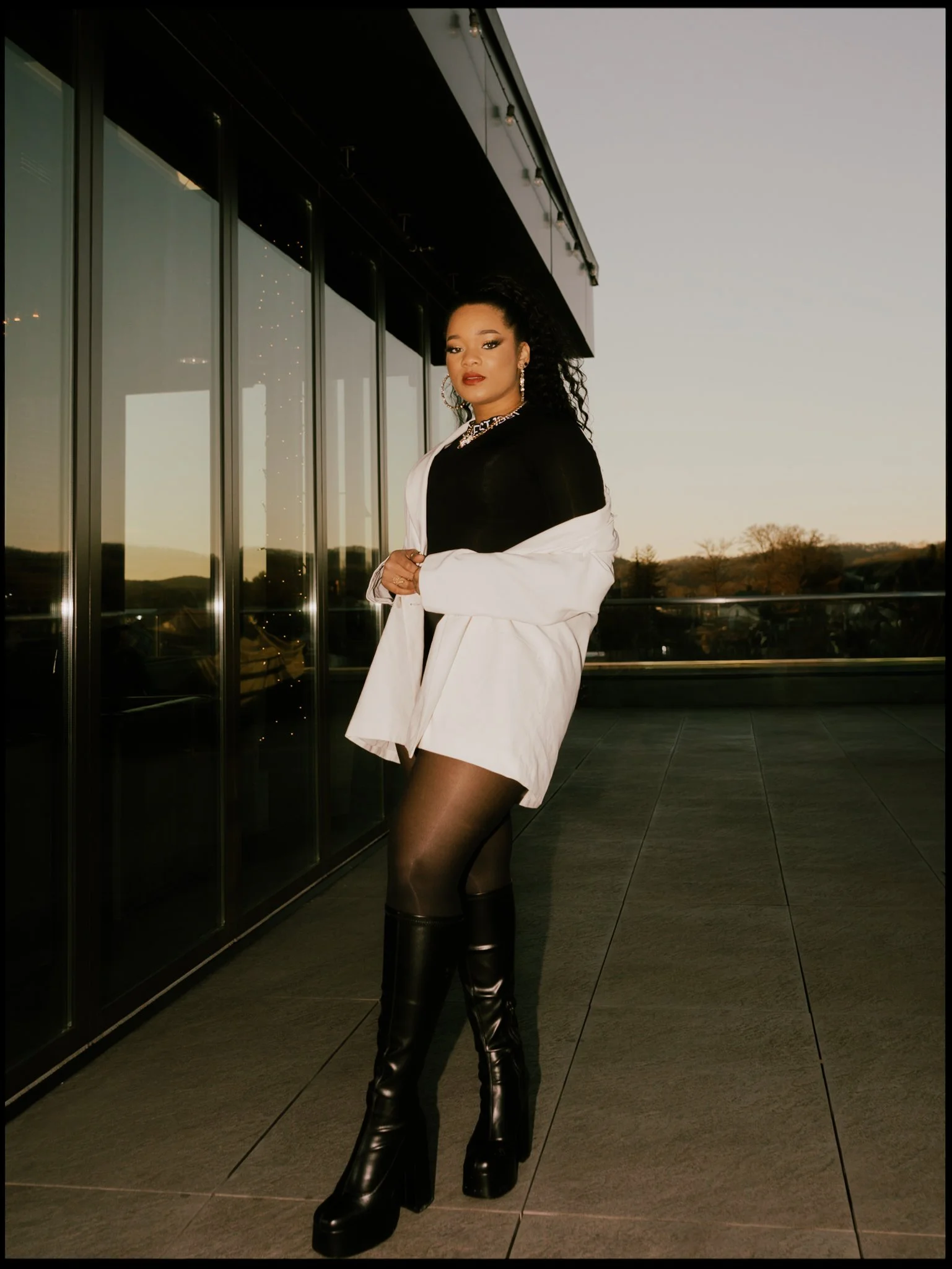 A woman standing on the street at night, wearing a black top, white blazer, and black thigh-high platform boots, with large earrings and long curly hair.
