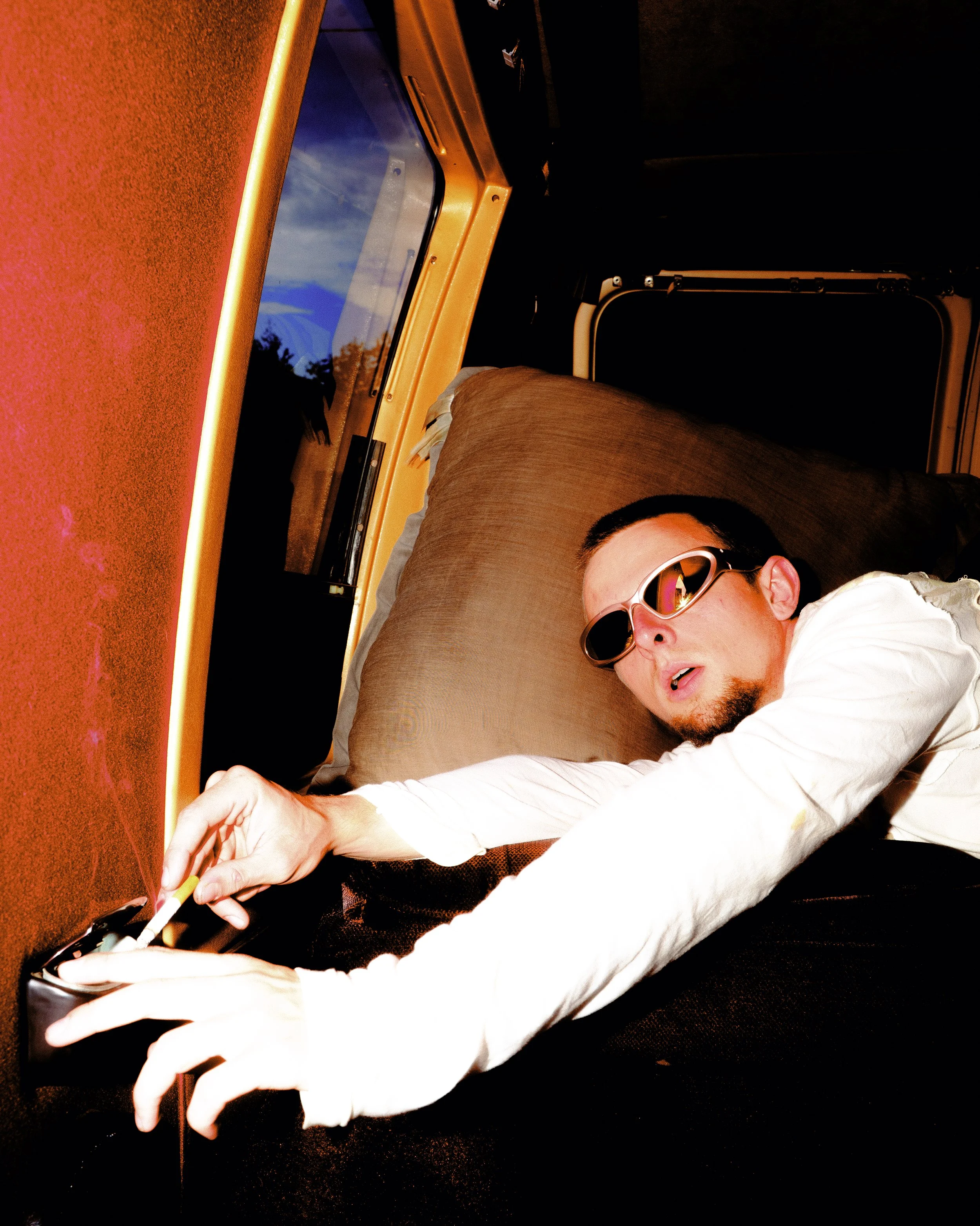 A man wearing sunglasses lying down inside a vehicle, reaching for a device on a table, with a window showing a blue sky and trees outside.