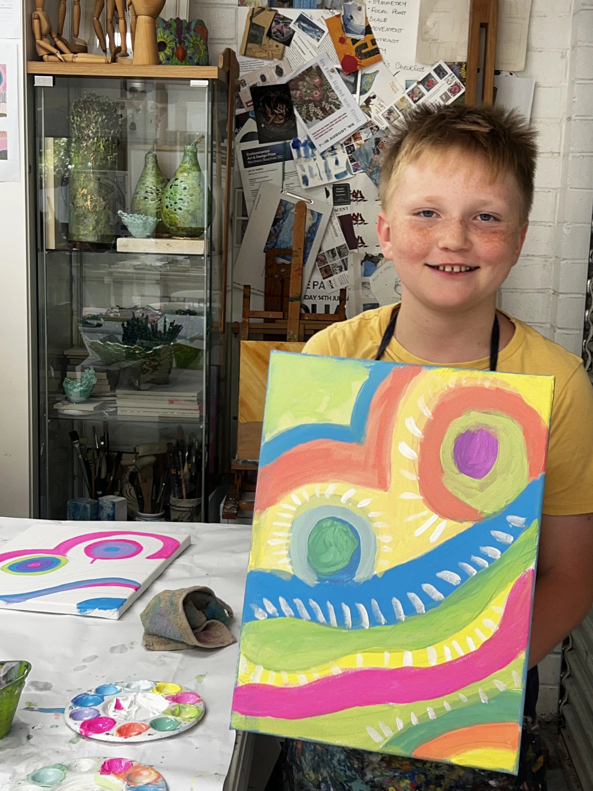 A young boy with red hair and freckles smiling while holding a colorful abstract painting. The painting features swirling circular shapes and vibrant colors like pink, yellow, blue, green, and purple. On the table in front of him are painting supplies, including a palette with pink, blue, green, and yellow paints, and another artwork with concentric circles. The background includes a glass cabinet with various pottery and decorations, a cluttered bulletin board with photos and papers, and white brick walls.