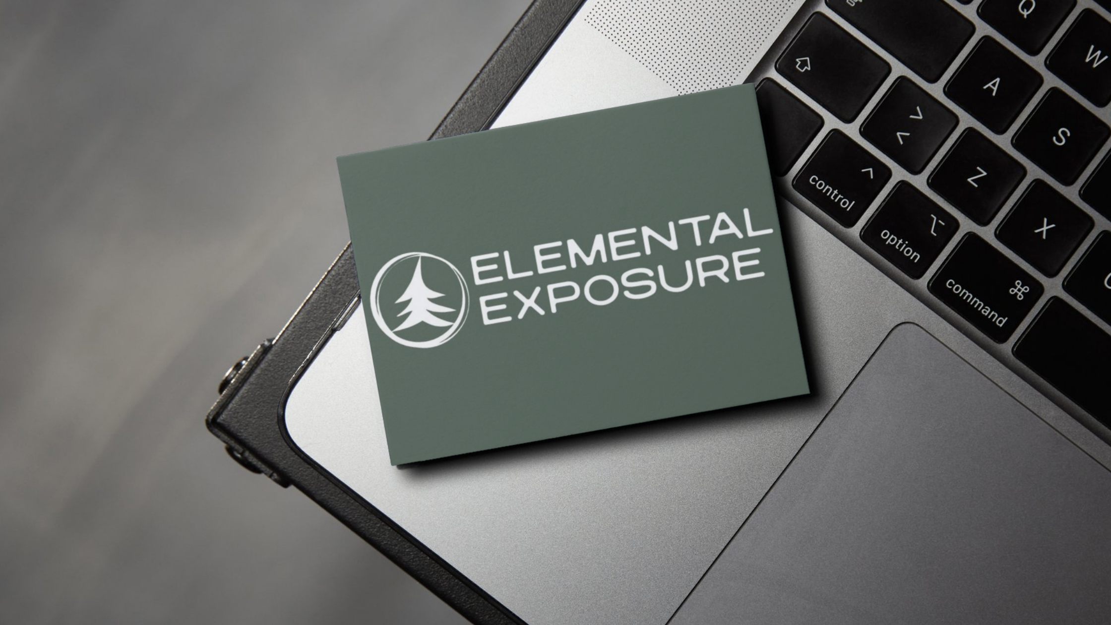 Business card mockup featuring Elemental Exposure logo with circular tree icon placed on a green card sitting on a laptop keyboard