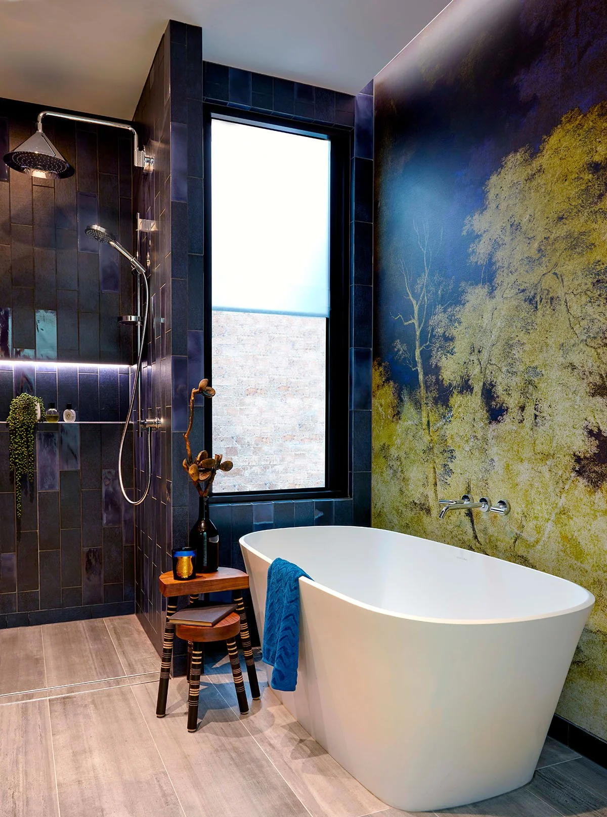 Modern bathroom with a white freestanding bathtub, a large window, a dark tiled shower area with a rain showerhead and handheld shower, a small wooden stool with a black vase holding twigs, and a colorful wall mural of a tree on a blue and yellow bac
