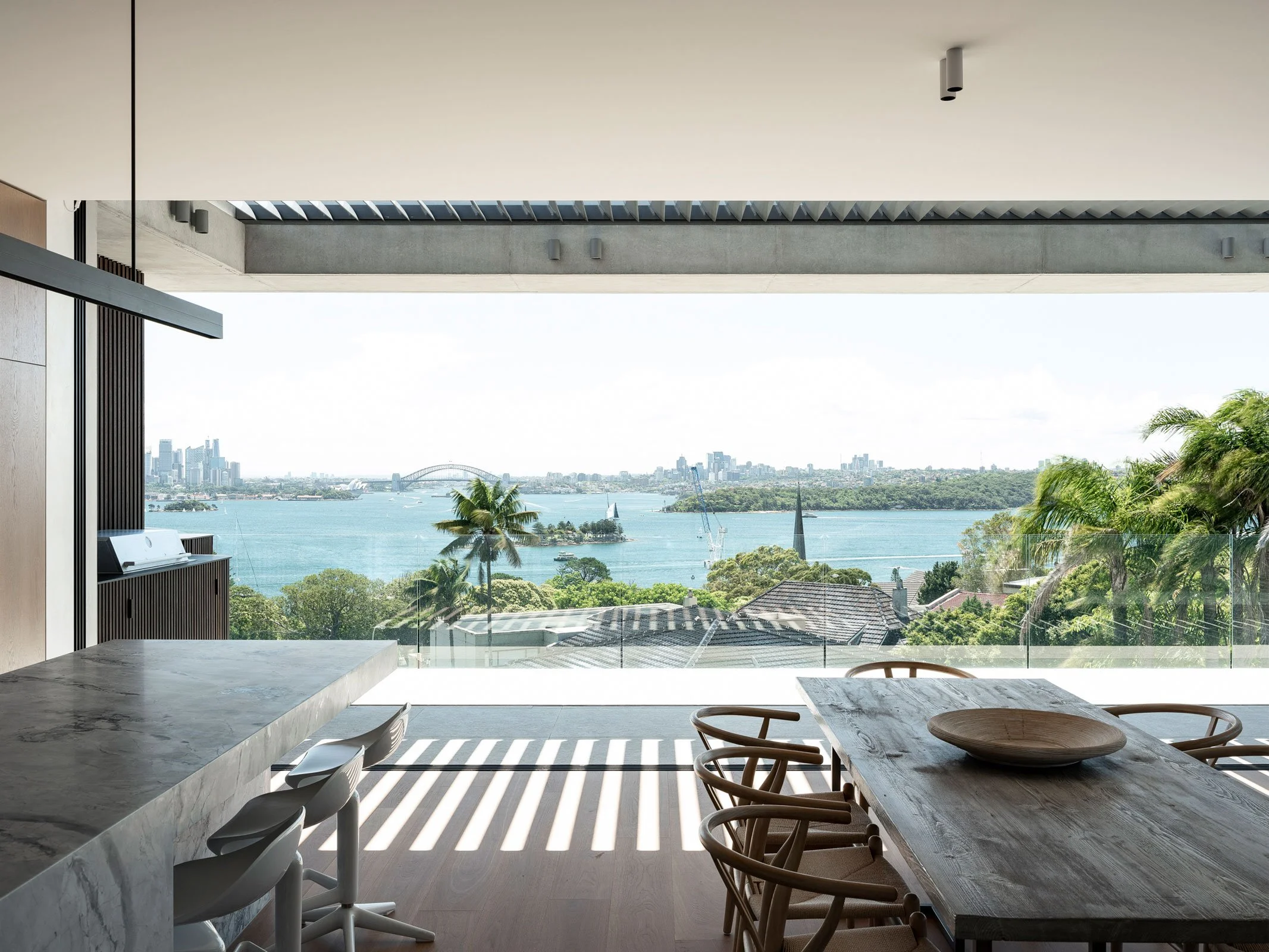 Modern dining area with a view of a river, city skyline, and greenery through large windows, with a wooden dining table and chairs, and a marble kitchen counter.
