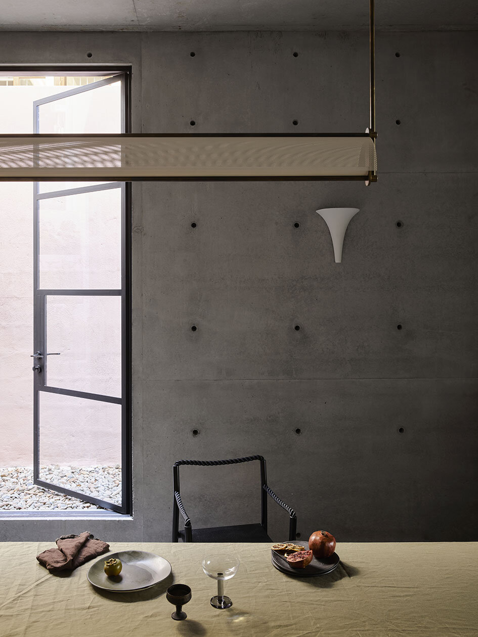 Interior of a modern room with concrete walls, a large open window, and a table set with a plate, a bowl, a glass, and pomegranate, with a chair and a decorative wall light.
