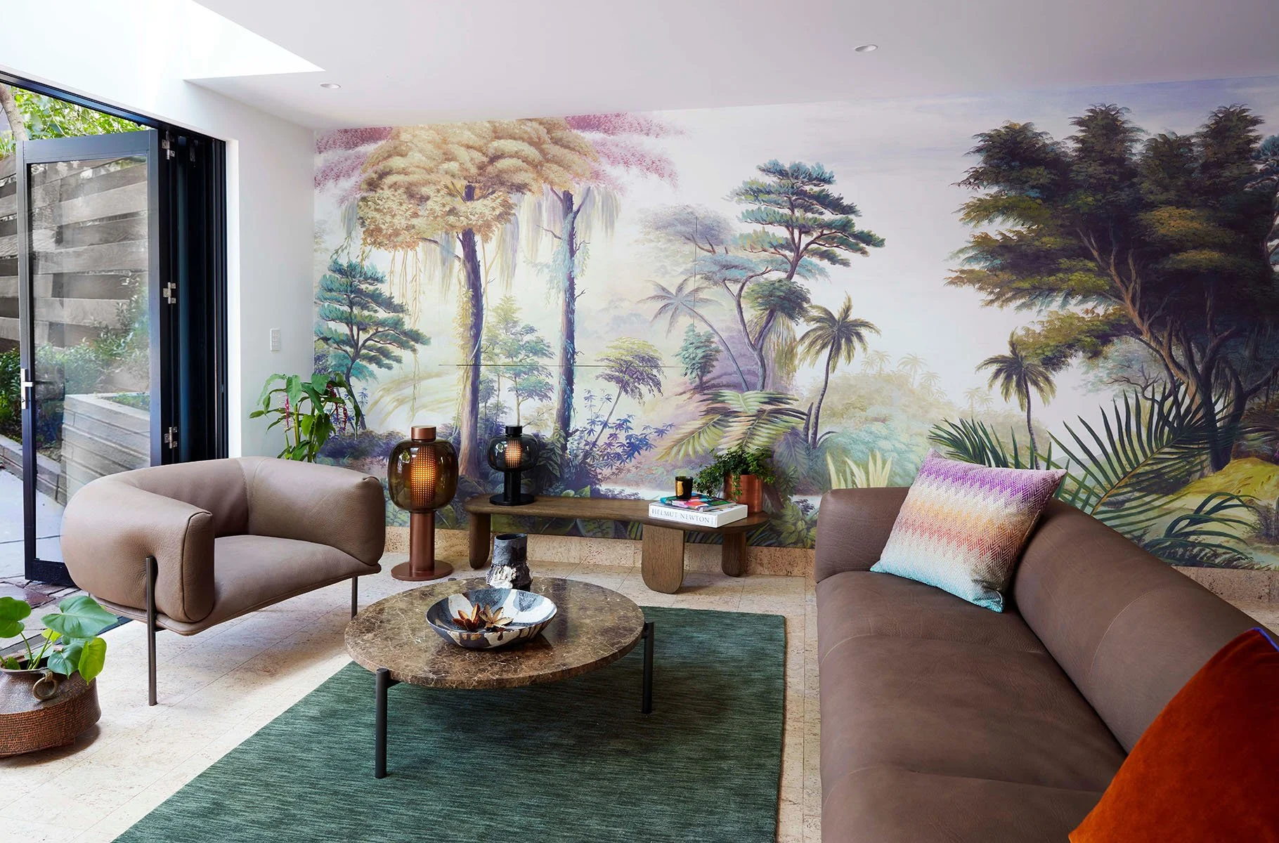 Living room with a mural of a forest landscape on the wall, tan armchair, brown sofa with colorful pillow, green rug, coffee table, potted plant, decorative vases, and a sliding glass door leading outside.
