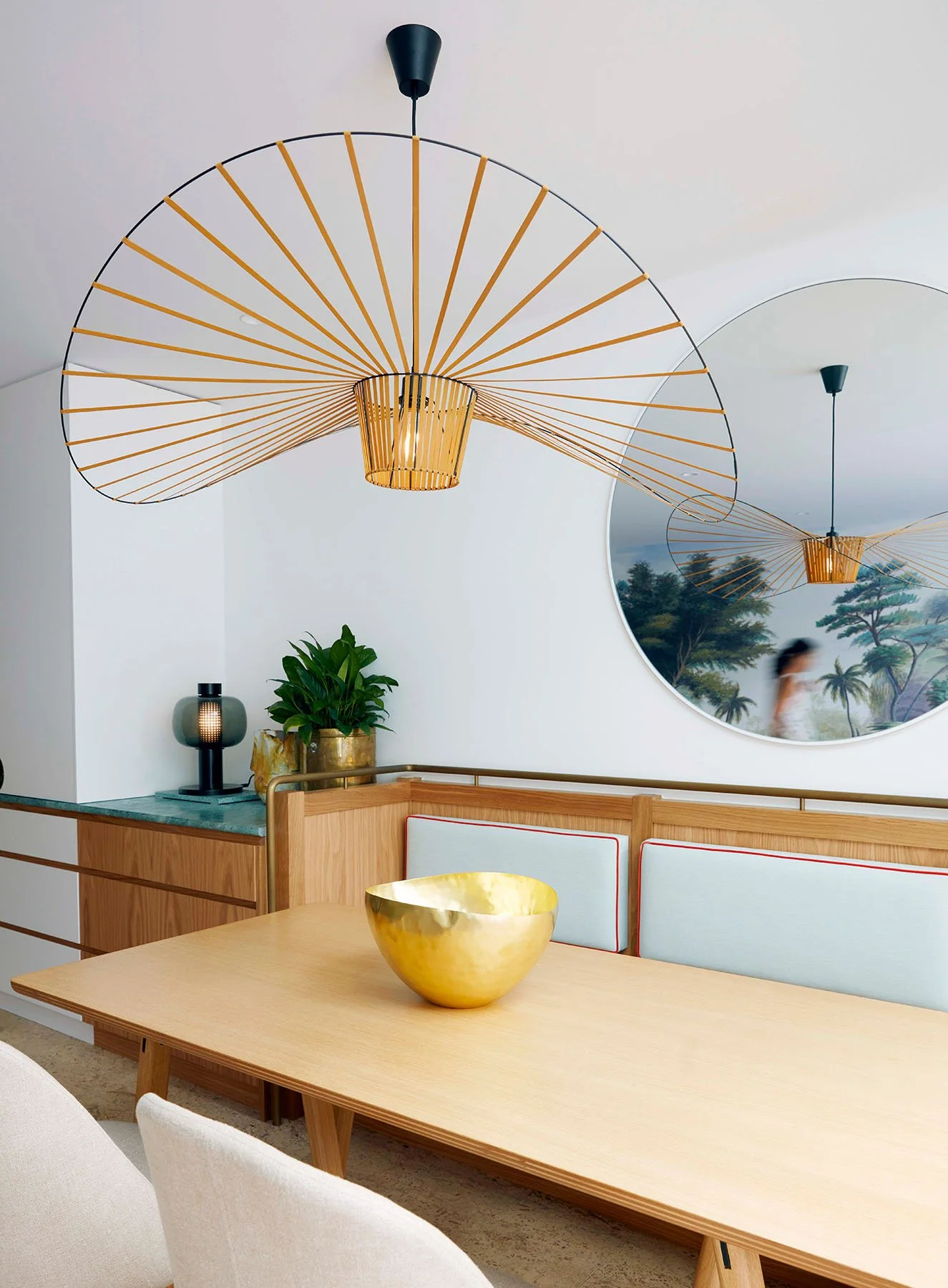 Interior dining space with light wood table, white chairs, gold bowl on table, wood-paneled wall with black lamp, green plant, large round mirror reflecting modern hanging light fixtures, and a door or window with a mural of trees and palm trees in t