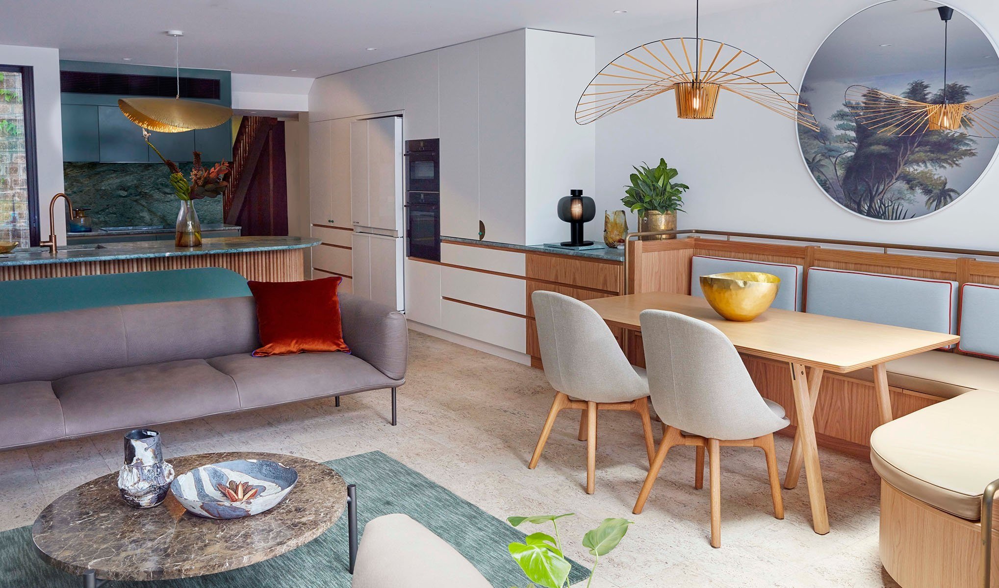 Modern open-concept living and dining area with a beige sofa featuring a red cushion, a round marble coffee table with decorative bowls, a dining table with gray chairs, and a built-in sideboard with plants and decorative items. The space has artwork