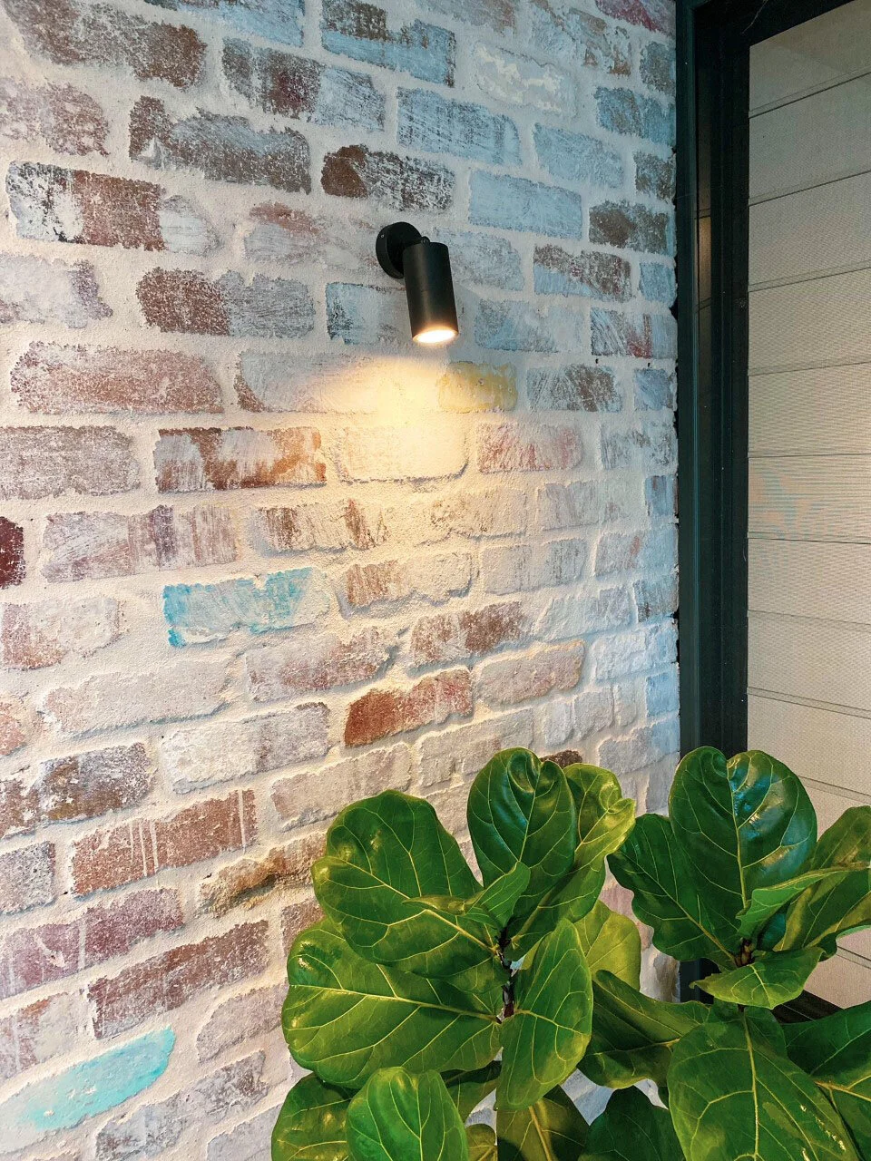 A black outdoor wall light mounted on a multicolored brick wall, illuminating the wall, with green leafy plants at the bottom next to a corner of a building.