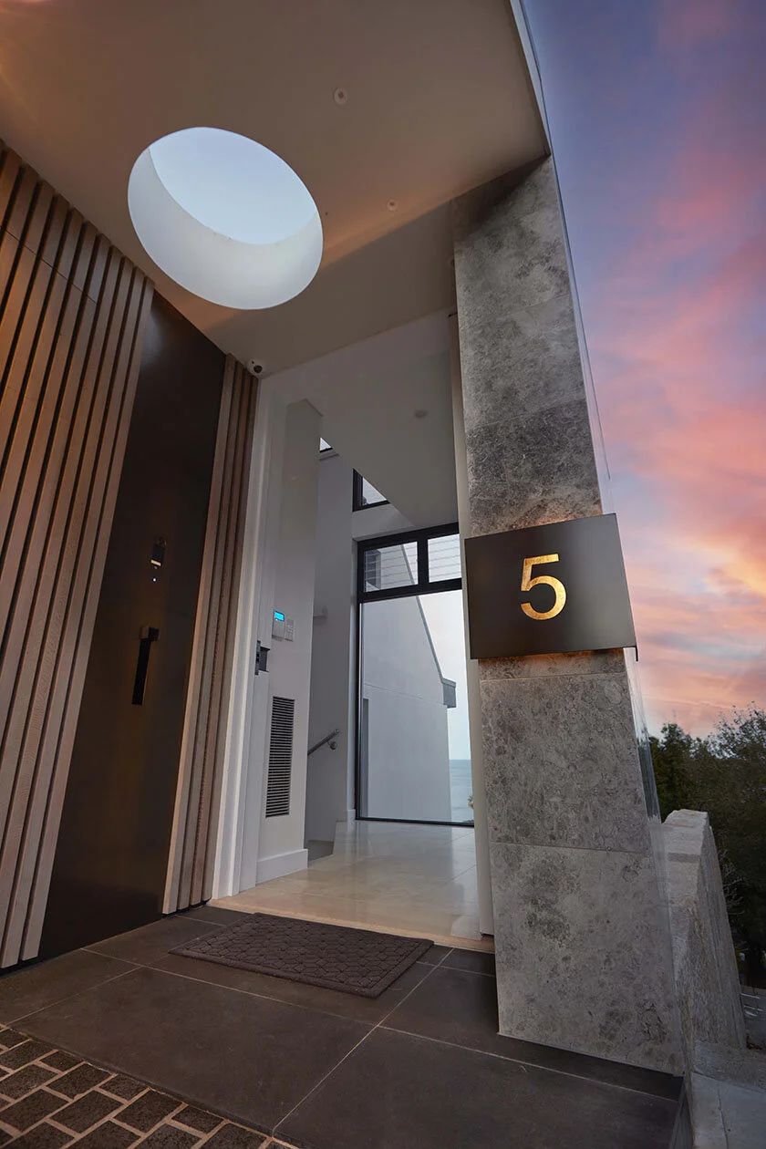 Apartment entrance with modern design, number 5 on a sign, large window showing a sunset sky, and interior features including wooden slats and a textured wall.