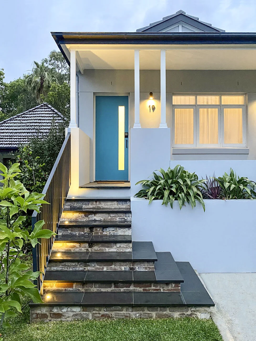 Front view of a modern house with a blue front door, illuminated staircase, and landscaped garden.