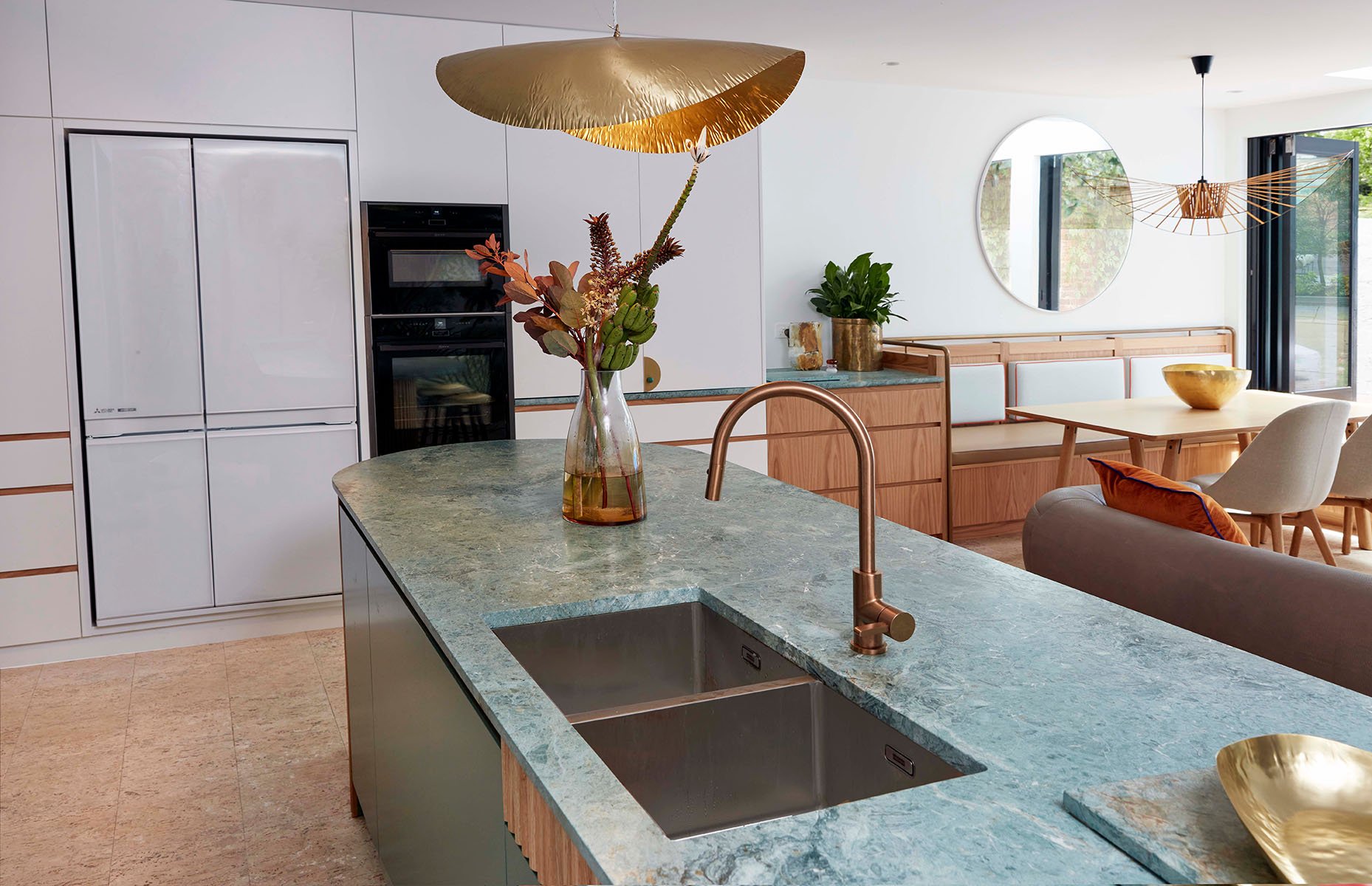 Modern kitchen with green marble island, brass fixtures, and wooden accents. Featuring a vase with foliage, built-in oven, refrigerator, dining area with bench and chairs, decorative bowls, and large windows with open doors.
