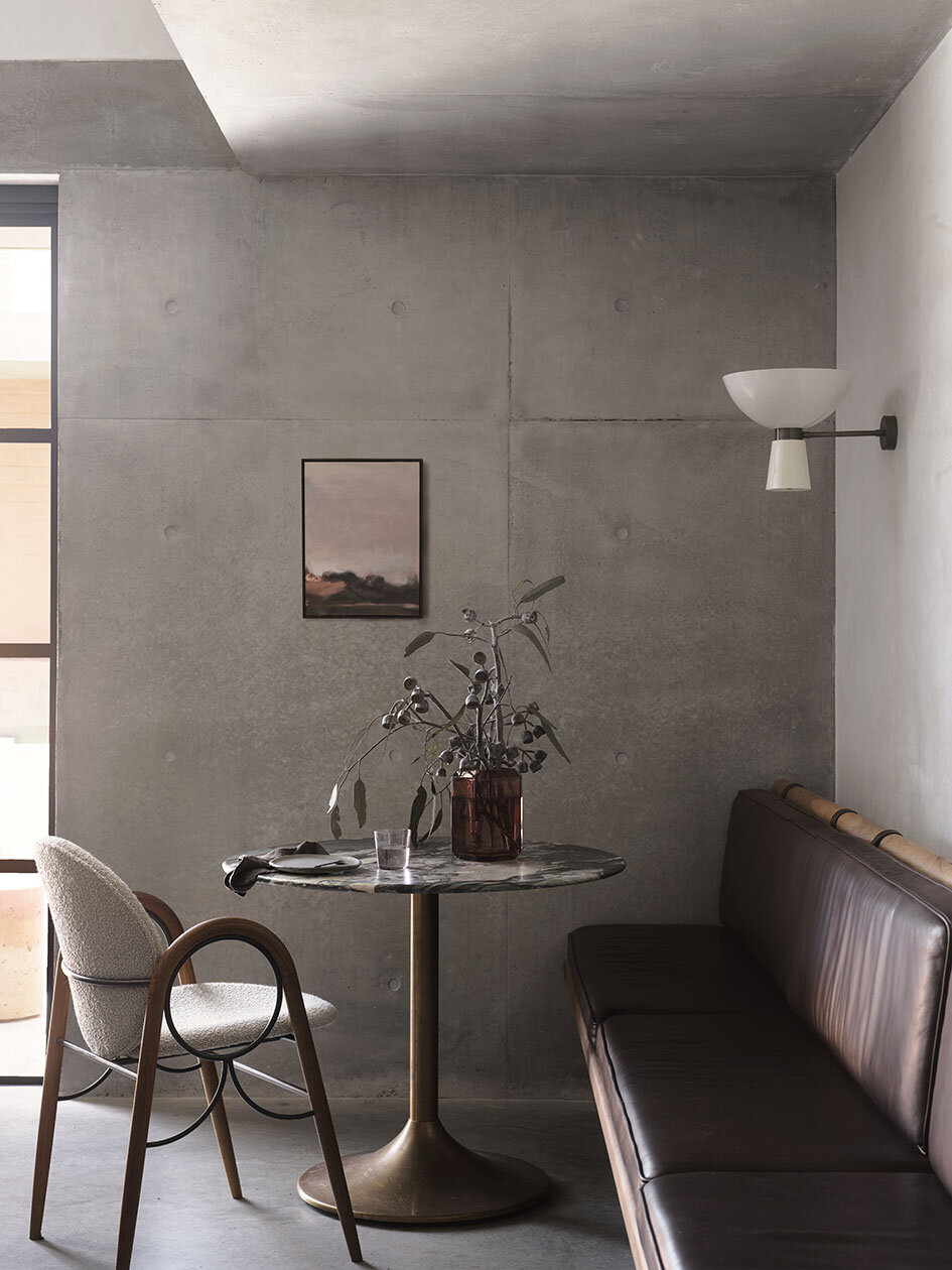 A minimalist interior with a concrete wall, a small round marble table, a gray upholstered chair, a brown leather bench, a glass vase with green branches, a glass of water, a small plate, a framed landscape painting, and an off-white wall-mounted lam