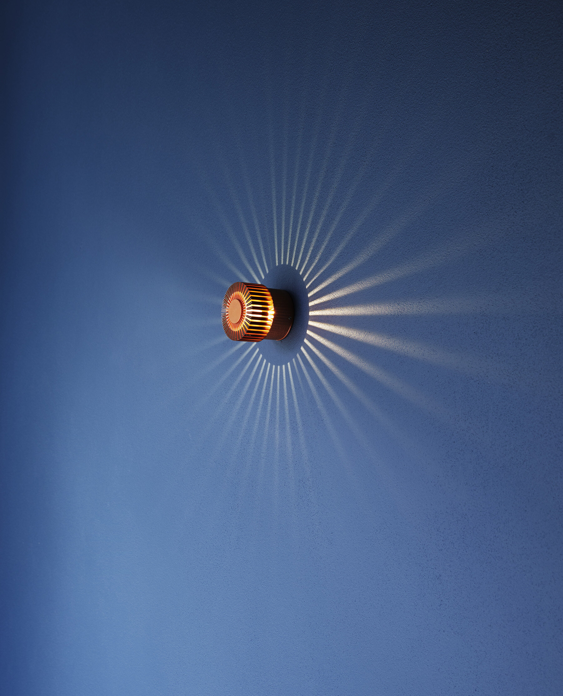 A modern wall-mounted light fixture with a circular design casts a starburst pattern of light and shadow on a blue wall.