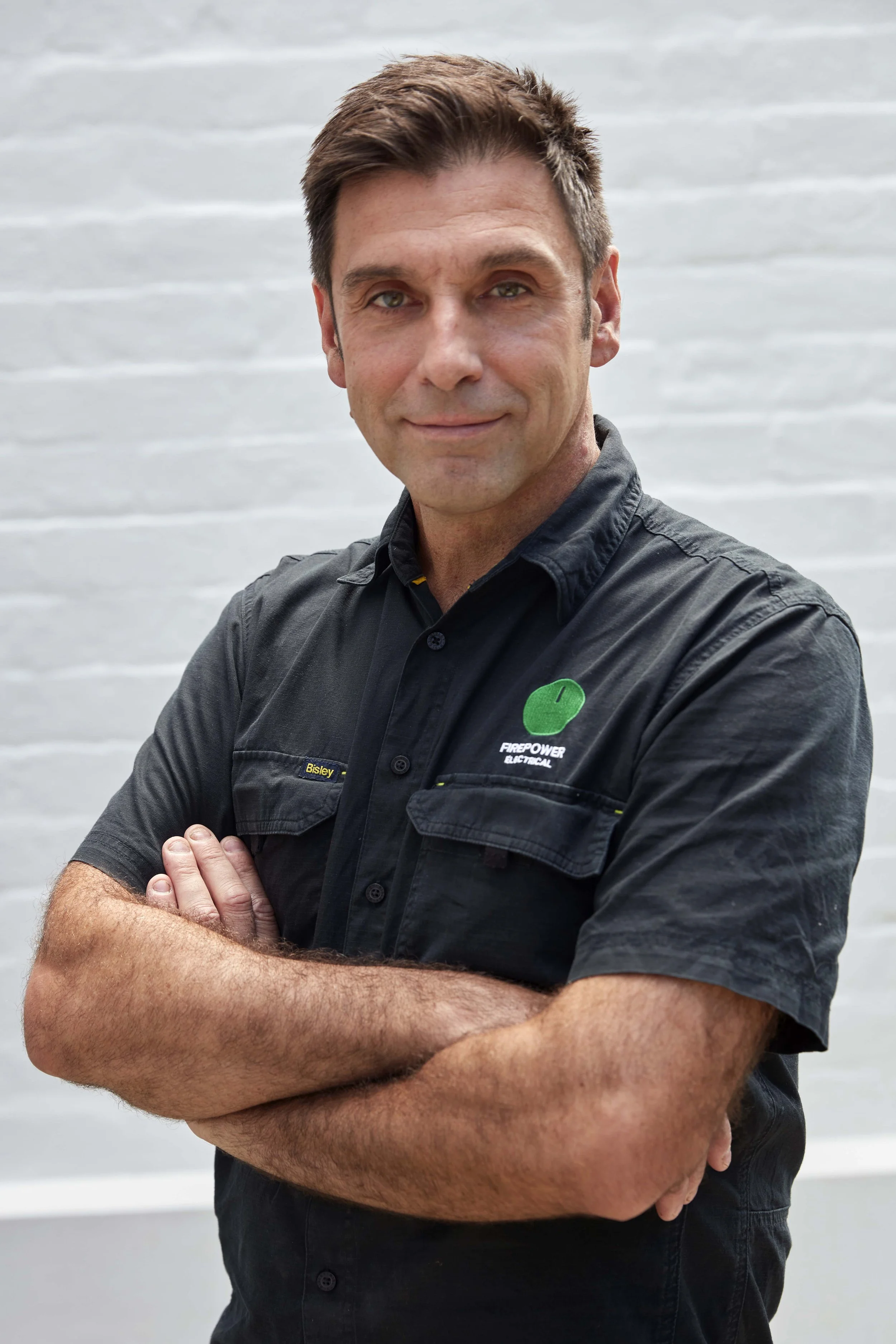 A man with short brown hair and light skin, wearing a black work shirt with a green logo and crossed arms, standing in front of a light-colored brick wall.