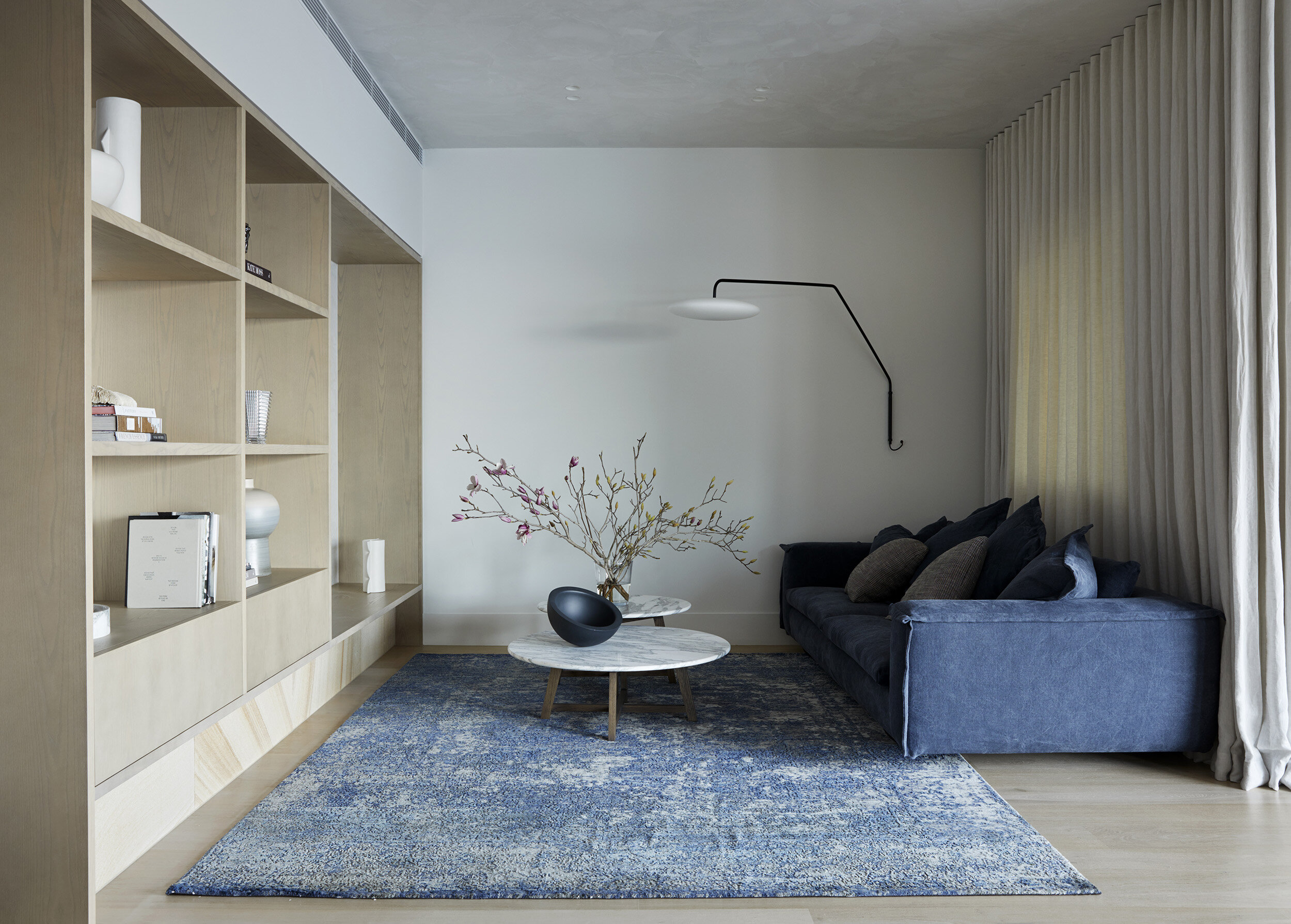 Modern living room with a blue velvet sofa, a white marble coffee table with a black bowl and flower arrangement, a built-in wooden bookshelf, a textured blue area rug, and a wall-mounted black and white reading light, alongside floor-to-ceiling curt