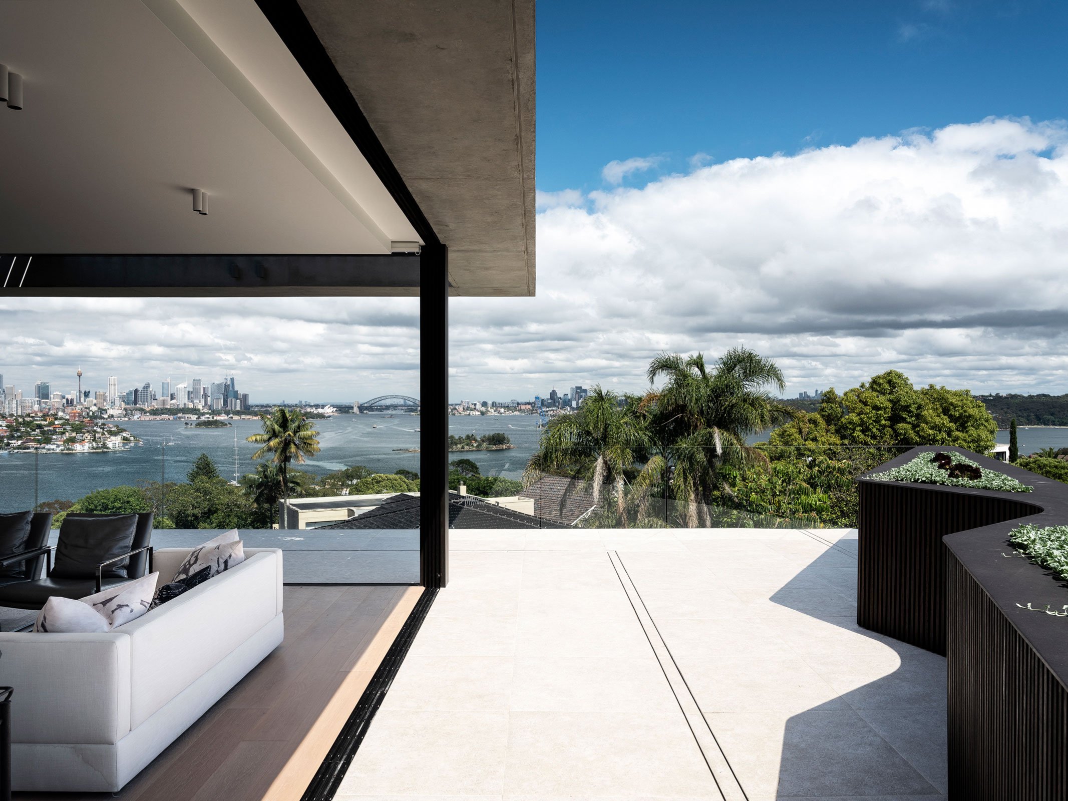 Modern balcony with white sofas and black chairs, overlooking a city skyline and water with palm trees and greenery in the background under a partly cloudy sky.