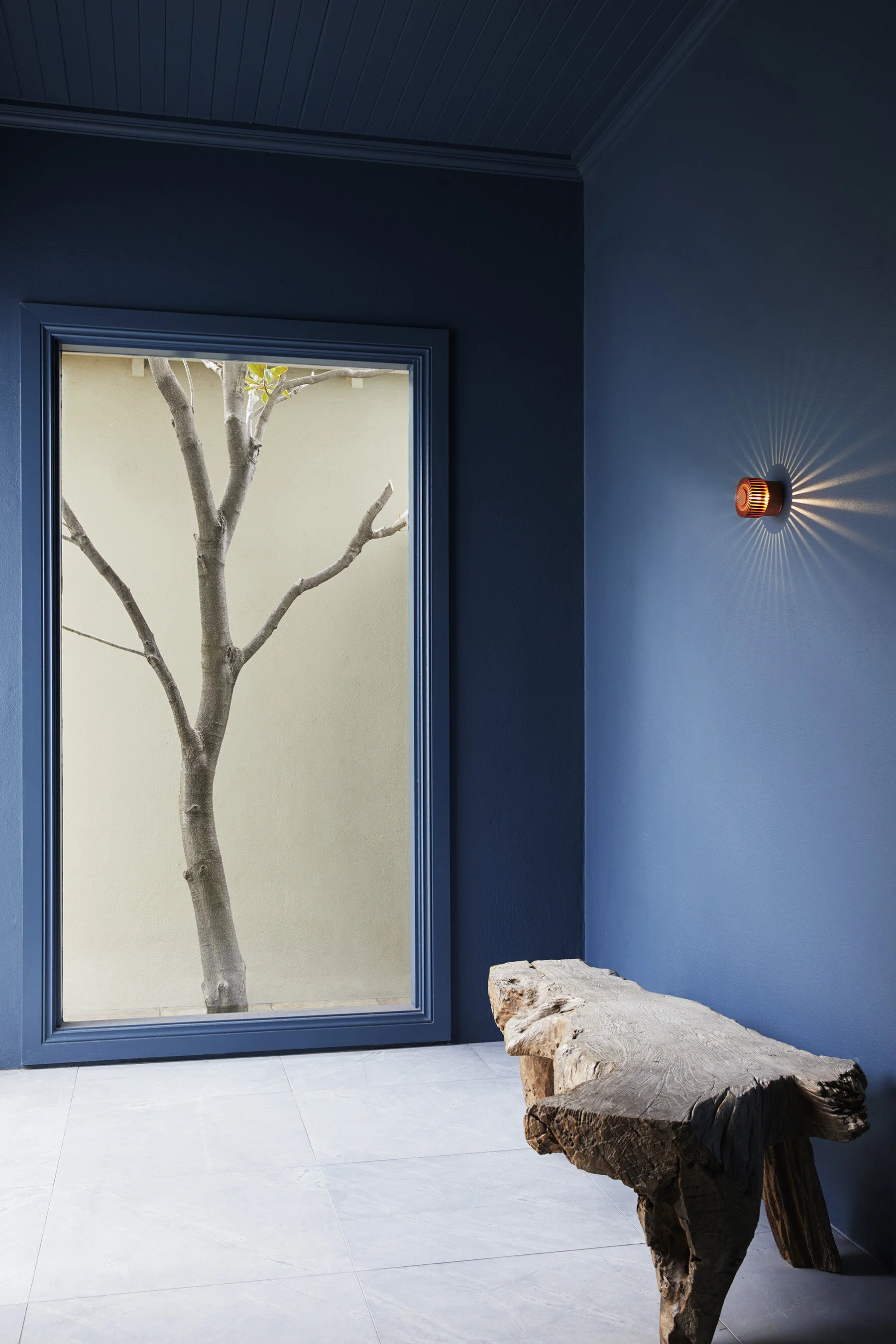 Interior with a blue wall, a window showing a tree outside, a wooden bench, and a wall-mounted orange light fixture.