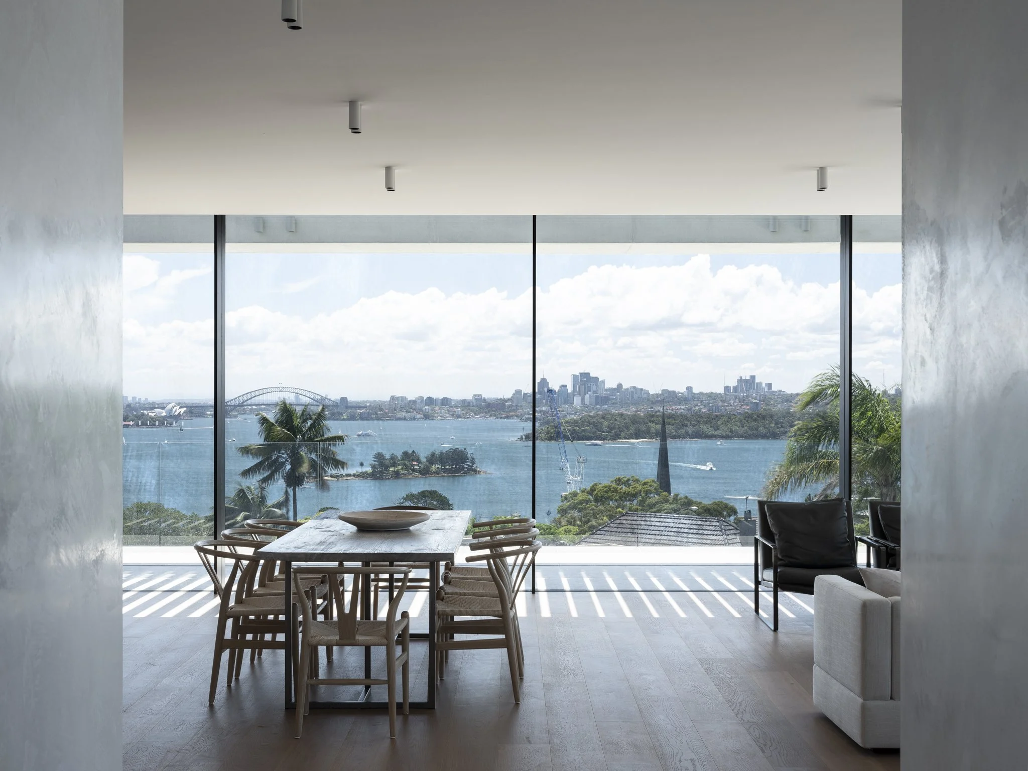 Modern living room with floor-to-ceiling windows overlooking a river, city skyline, and trees, with a dining table and chairs, chairs, and a sofa.
