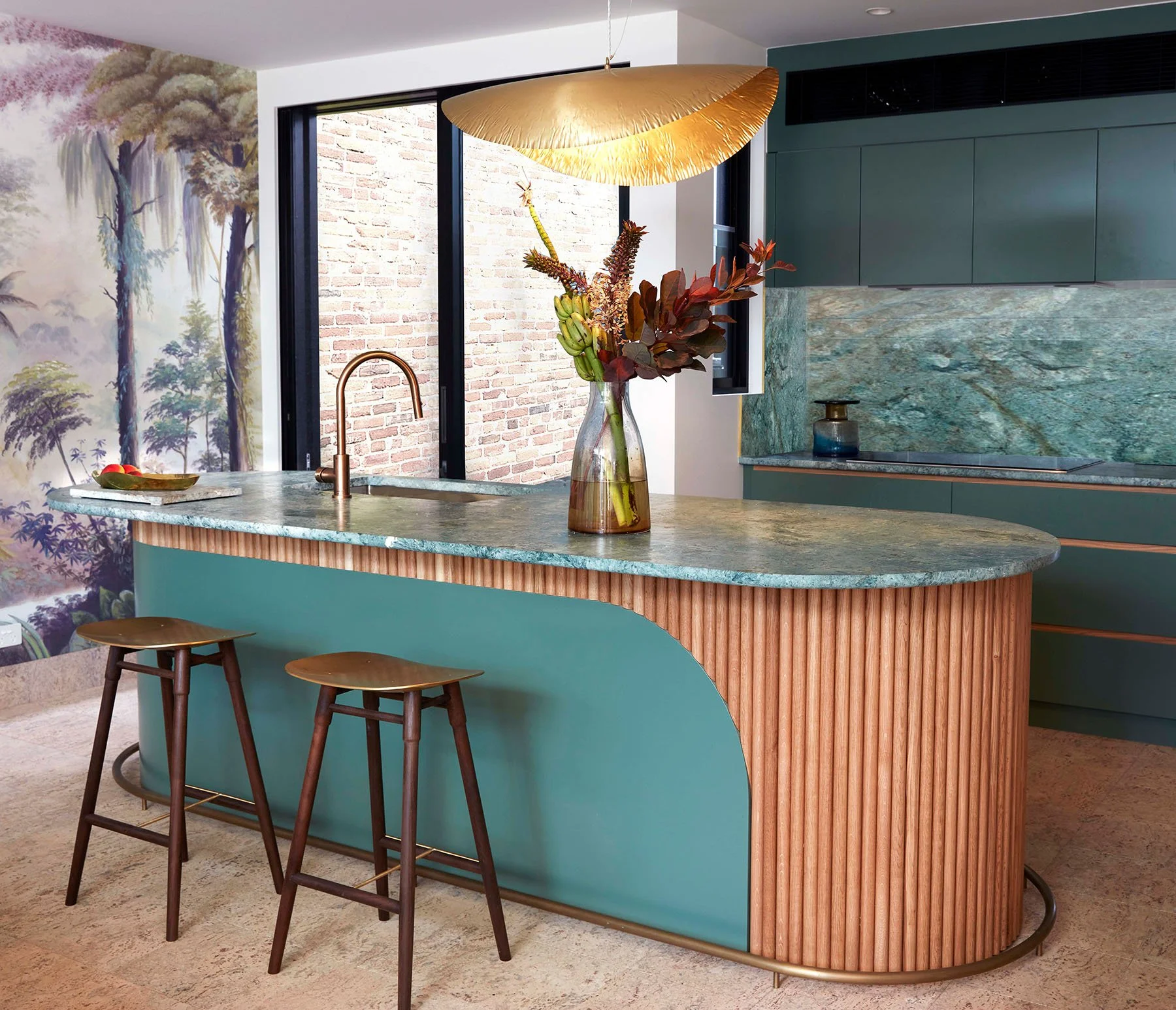 Modern kitchen with green tile countertops and cabinets, a curved kitchen island with wood paneling and green base, two wooden barstools, a large glass vase with dried flowers, and a decorative lighting fixture overhead. A large window reveals a bric