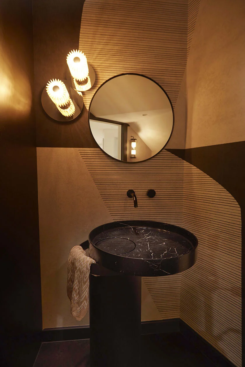 A modern bathroom with a black marble sink on a black pedestal, a round mirror above, and two decorative wall sconces with a sunburst design, warm lighting, and textured walls.