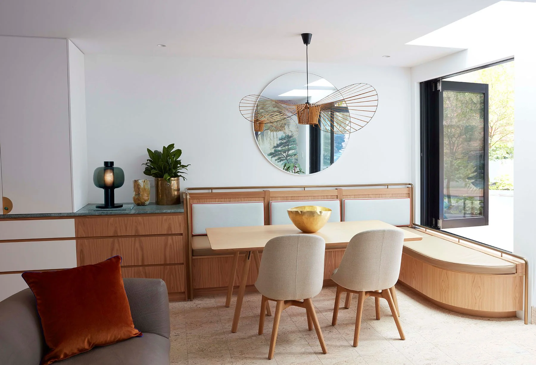 Minimalist dining area with a light wood table, two beige chairs, a built-in bench, decorative bowl, wall mirror, plants, and a large open window with a view of trees.