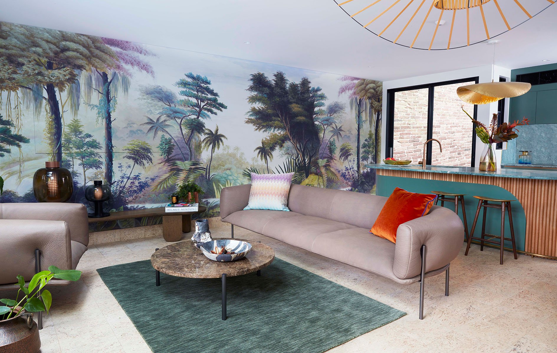 Living room with a mural of a lush jungle landscape on the wall, beige sofas with colorful cushions, a green rug, a marble coffee table, and a kitchen area with a turquoise counter and bar stools.