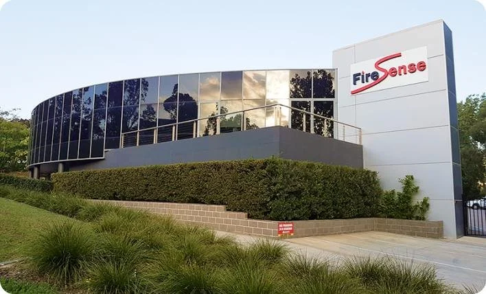 Exterior of a modern office building with large reflective glass windows and a sign that reads 'FireSense' near the entrance, surrounded by landscaped bushes and grass.
