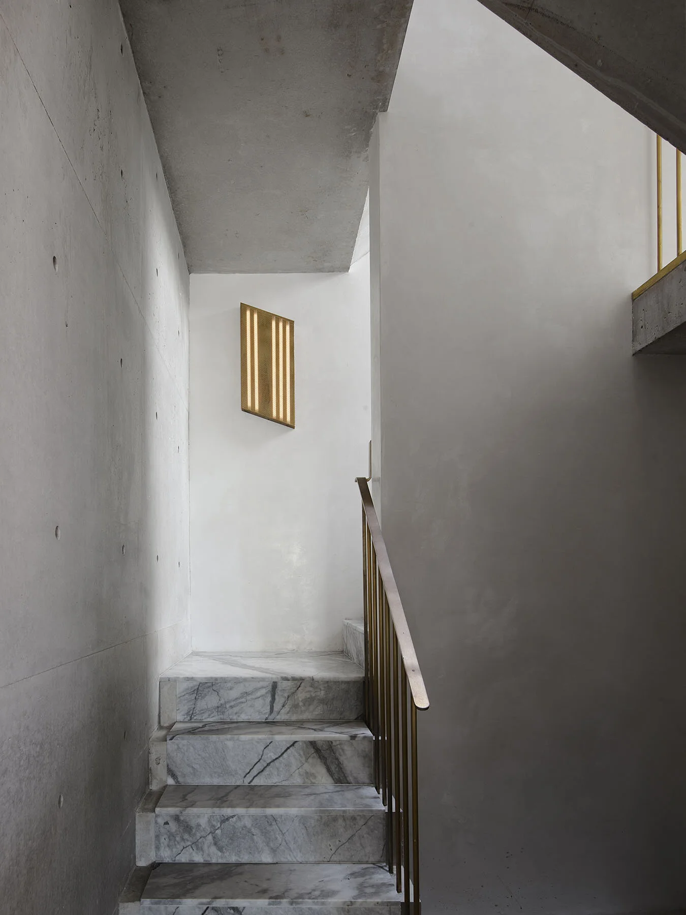 Modern staircase made of white marble with a metal railing, leading up to a landing with a gold-colored railing. The surrounding walls are plain and light-colored, with a single piece of geometric wall art in gold.