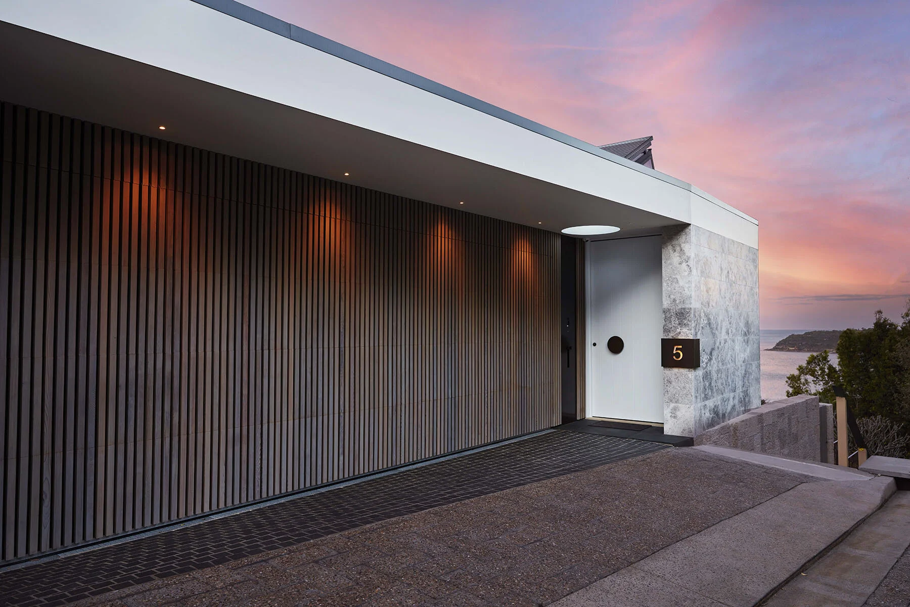 Modern house entrance with a white door, black circle handle, and house number 5 on a black plaque. The house features vertical wood paneling, gray stone accents, and a large overhanging roof. The background shows a scenic view of the ocean and a col