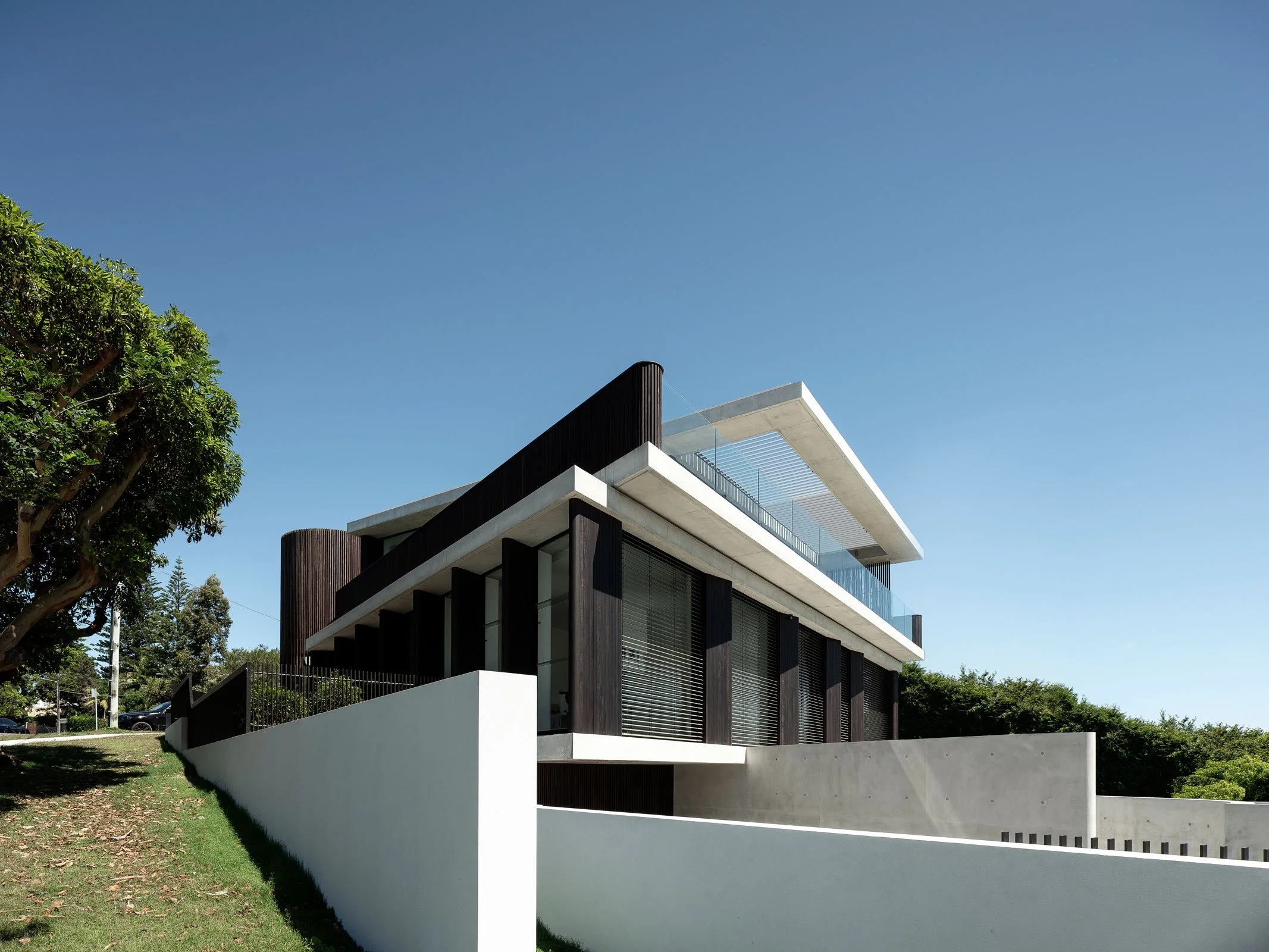 Modern multi-story house with white concrete walls, dark vertical wood panels, glass balcony, and flat roof, surrounded by green grass and trees under a clear blue sky.
