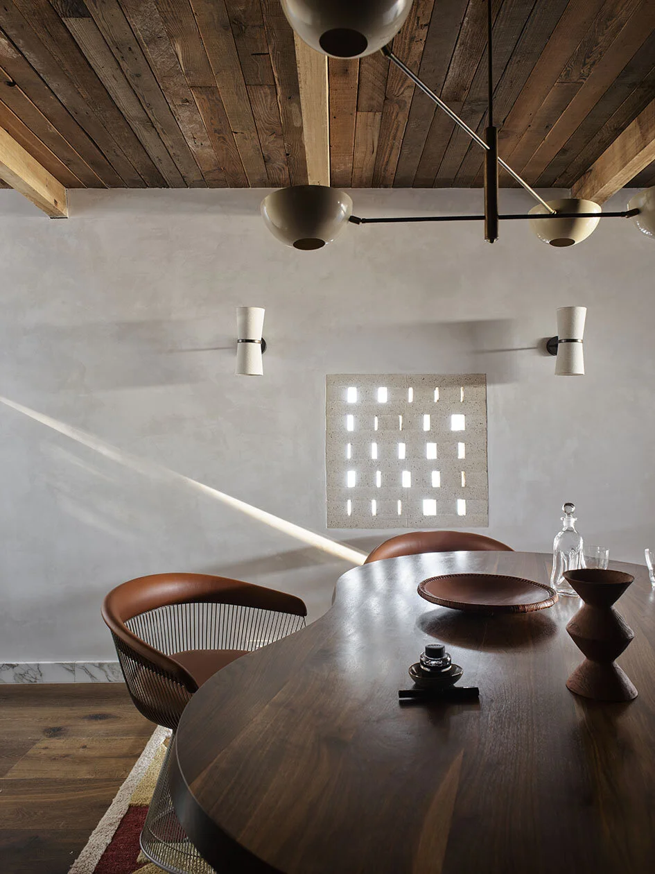 Interior of a modern dining room with a wooden table, brown chairs, a wall with small square openings letting light in, ceiling with wooden panels, and contemporary light fixtures.