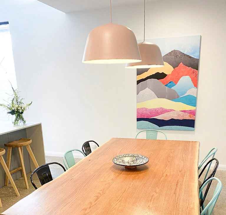 A modern dining room with a rectangular wooden table, six metal chairs, two beige pendant lamps hanging above, a colorful mountain landscape painting on the wall, and a white countertop with stools and a flower vase.