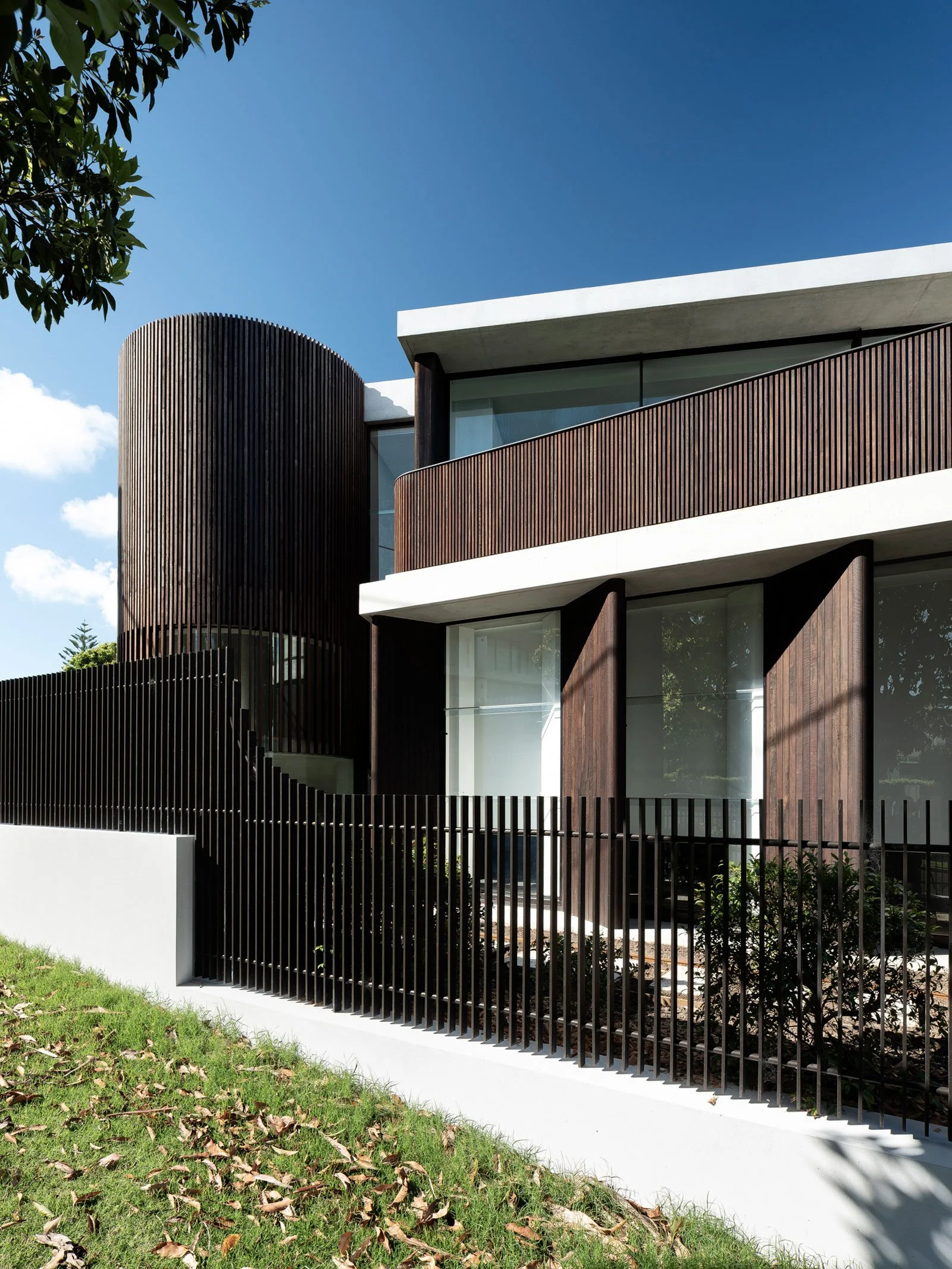 Modern house with curved wooden exterior walls, large glass windows, white balconies, and a black metal fence, under a blue sky with clouds.