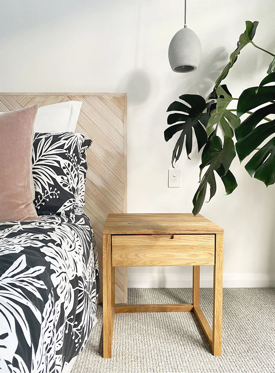 A bedroom with a wooden nightstand beside a bed, black and white patterned bedding, and pink and white pillows. A large green leafy plant is on the right, with a pendant light hanging from the ceiling and an electrical outlet on the wall.