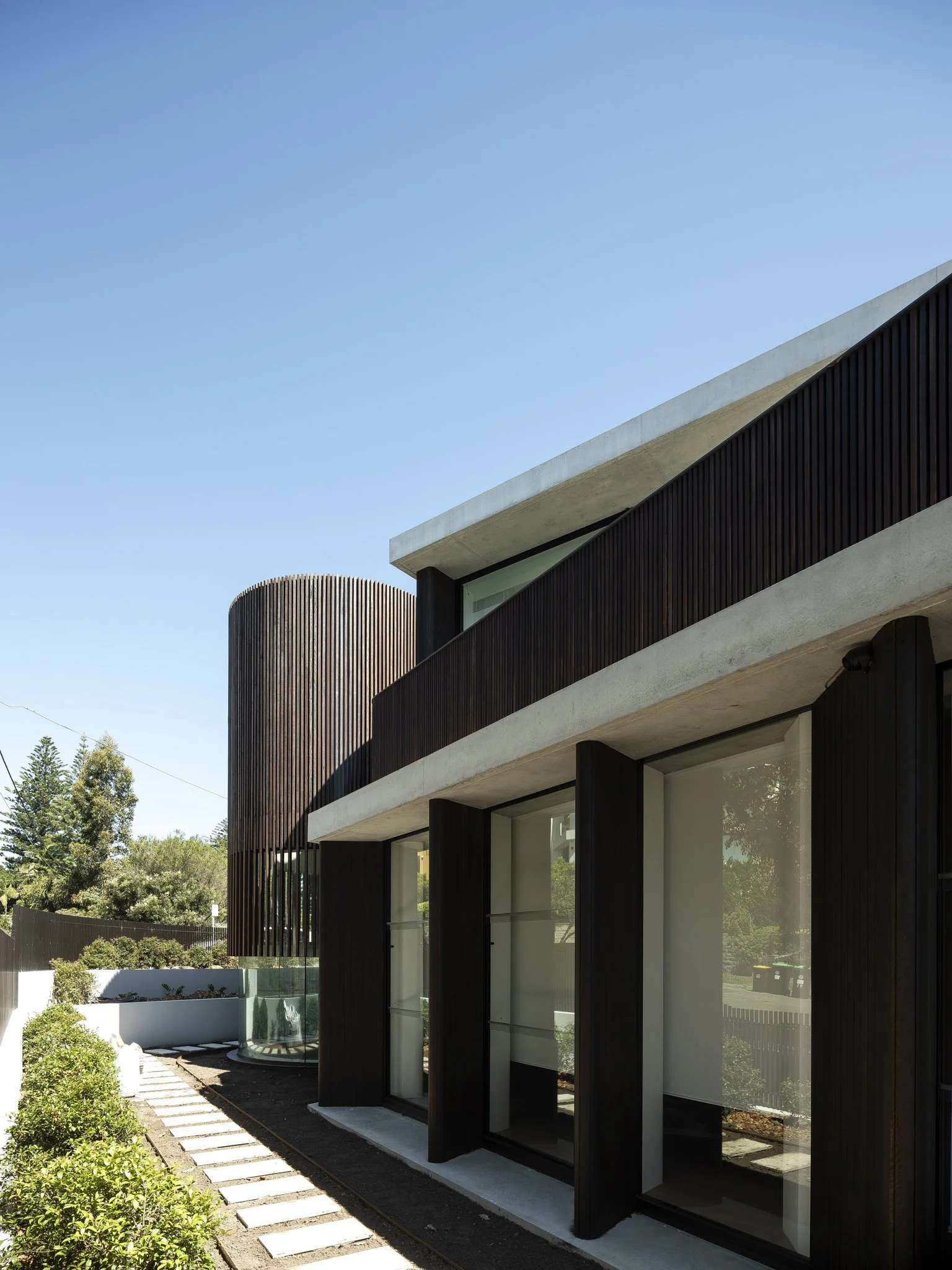 Modern building with dark wooden panels, large glass windows, and a curved vertical wood feature, set against a clear blue sky.