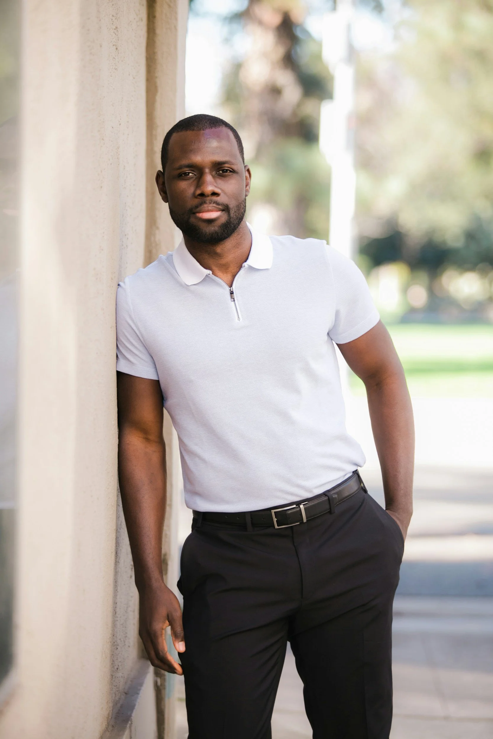 A man with short hair and a beard stands outdoors, leaning against a beige wall, wearing a white polo shirt and black pants, with a relaxed expression.