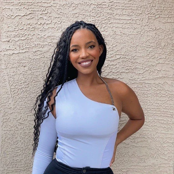 A smiling woman with long curly hair wearing a one-shoulder light blue top with a chain strap standing against a textured beige wall.